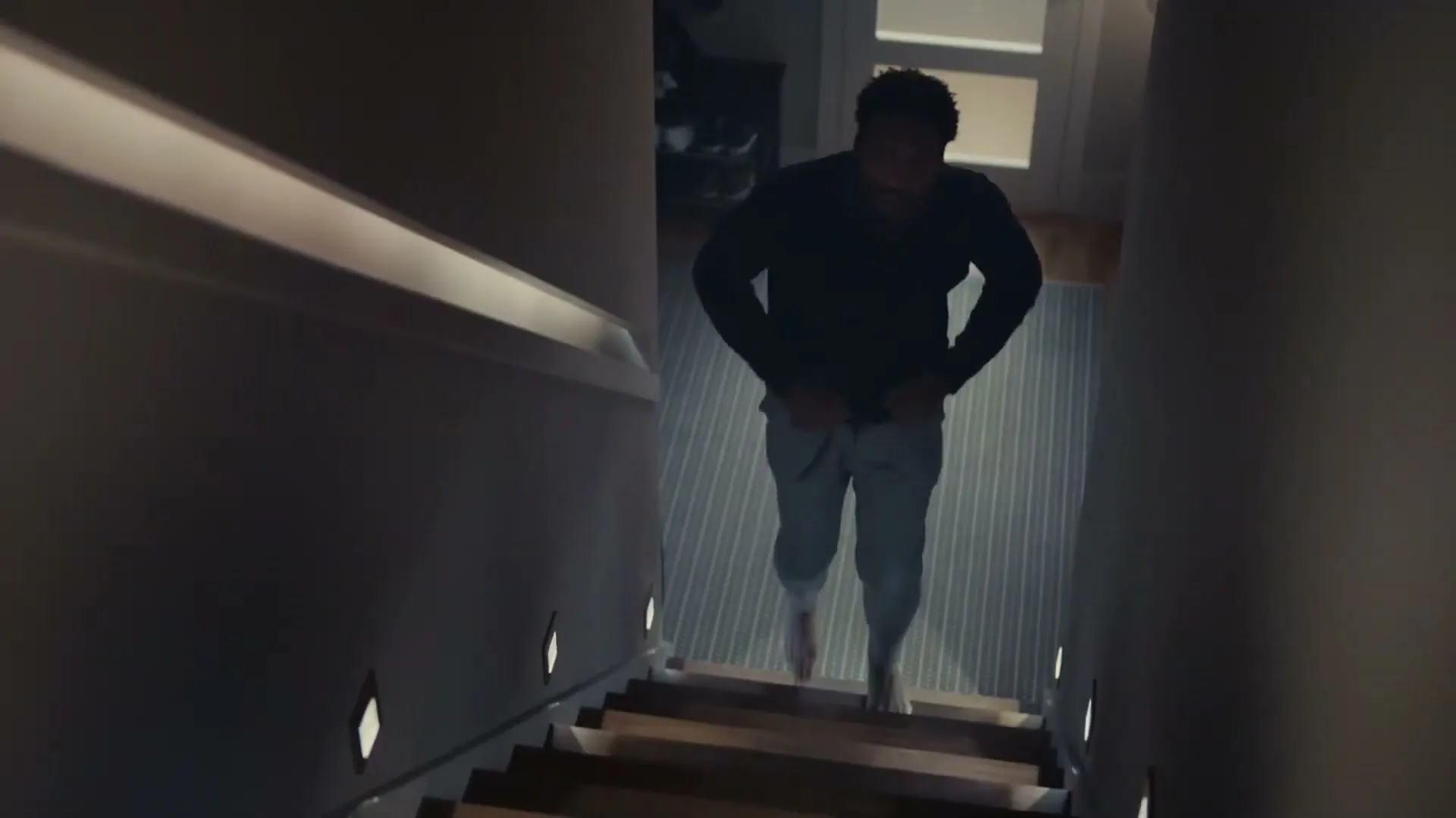 A man in his 30s, wearing a dark shirt and light pants, walks barefoot up a dimly lit staircase in a modern home at night.