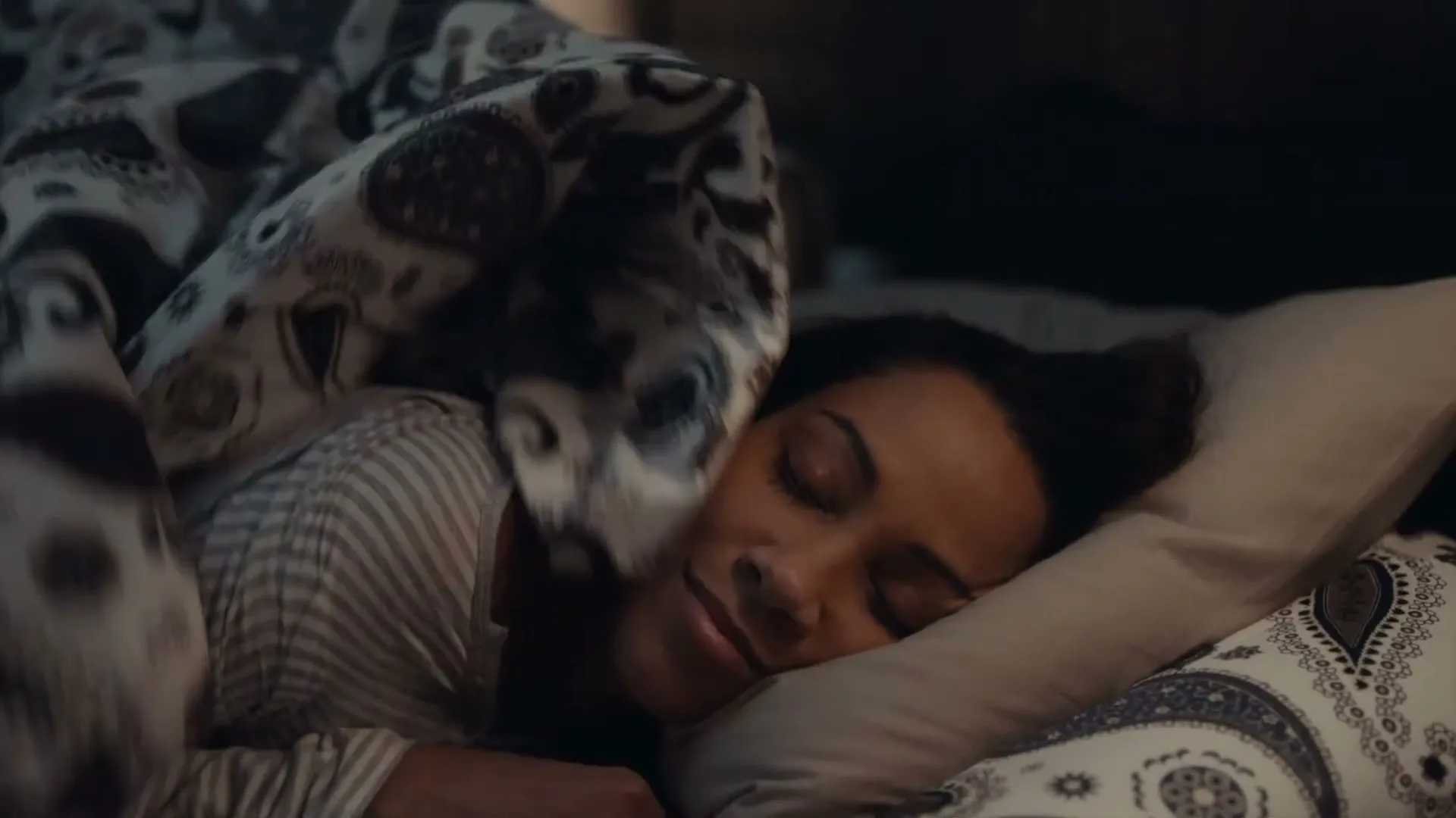A young woman with dark hair sleeps peacefully in bed, covered by a patterned duvet, in a dimly lit bedroom.