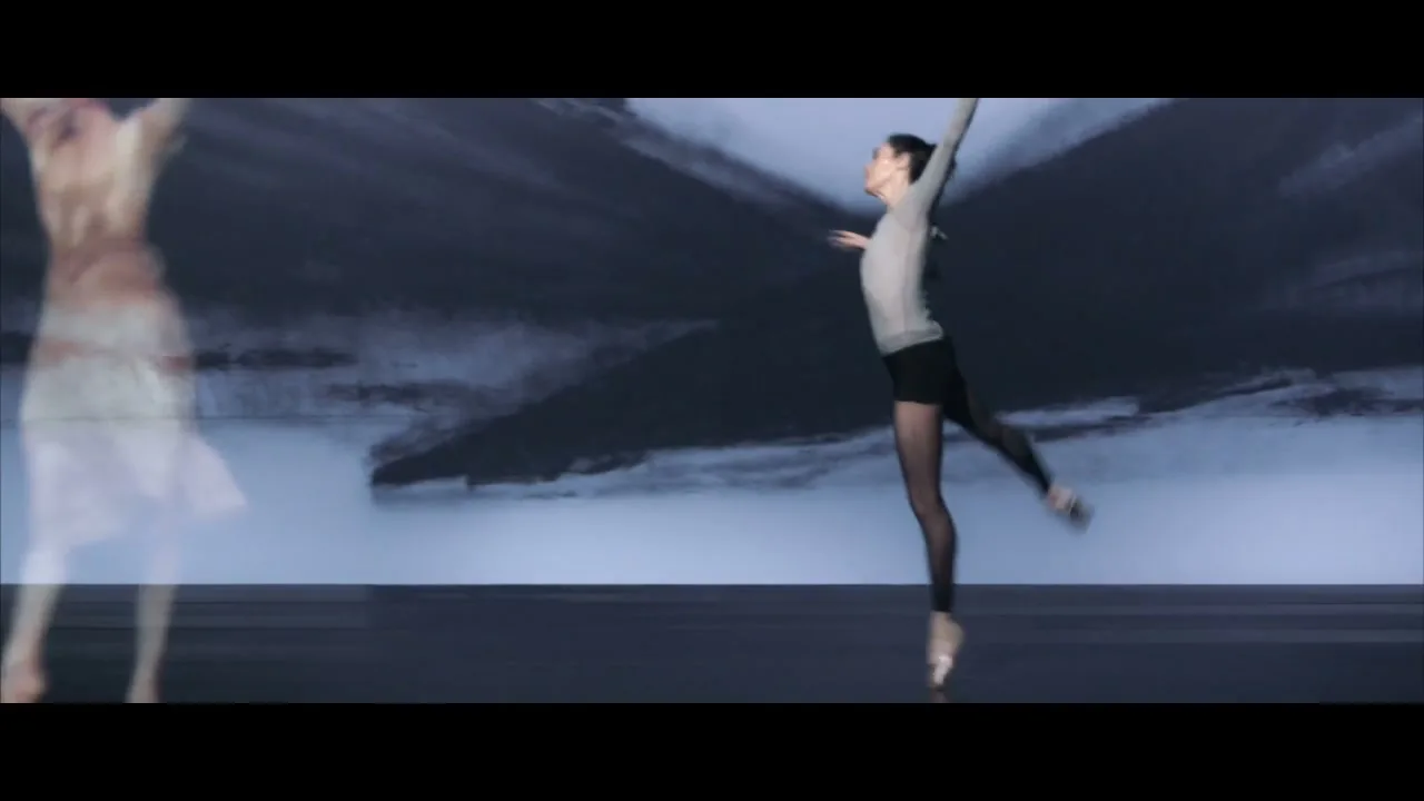 A female ballet dancer in a grey top and black shorts performs on a dark stage with a light blue and grey abstract background.