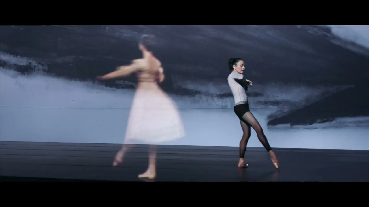 A female ballet dancer in a white top and black tights performs on a stage with a projected image of another dancer, in a studio.