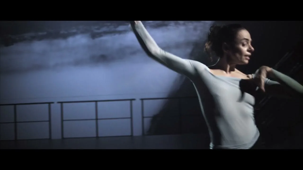 A female dancer in a light grey top performs a ballet move with arms outstretched in a dark studio with a cloudy projection.