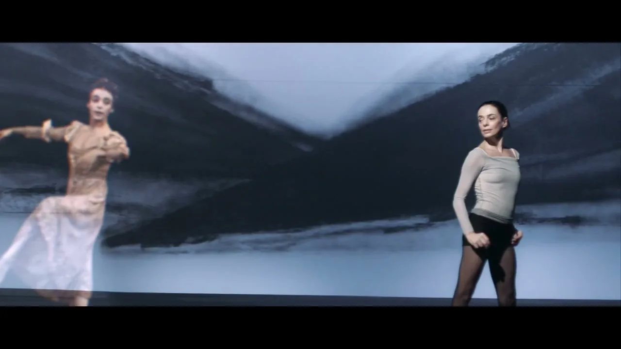 A female ballet dancer in a leotard stands on a stage with a projected image of a historical dancer and abstract art behind her.