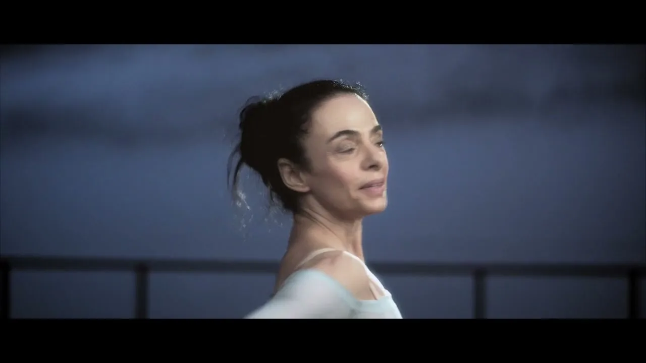 A female ballet dancer in her 40s, wearing a light blue leotard, turns and raises her arms in a dance studio with a dark blue background.