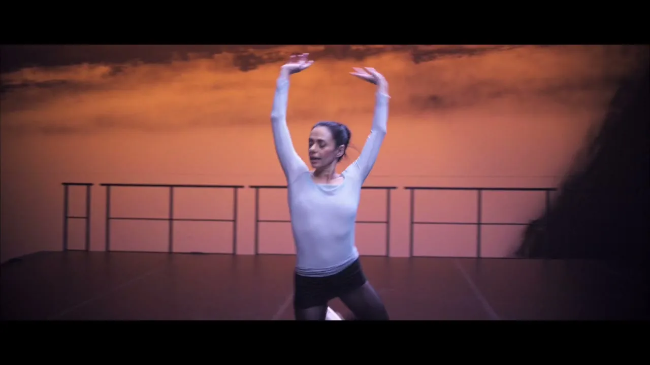A female dancer in a light top and dark shorts performs ballet moves on a stage with an orange, cloudy backdrop.