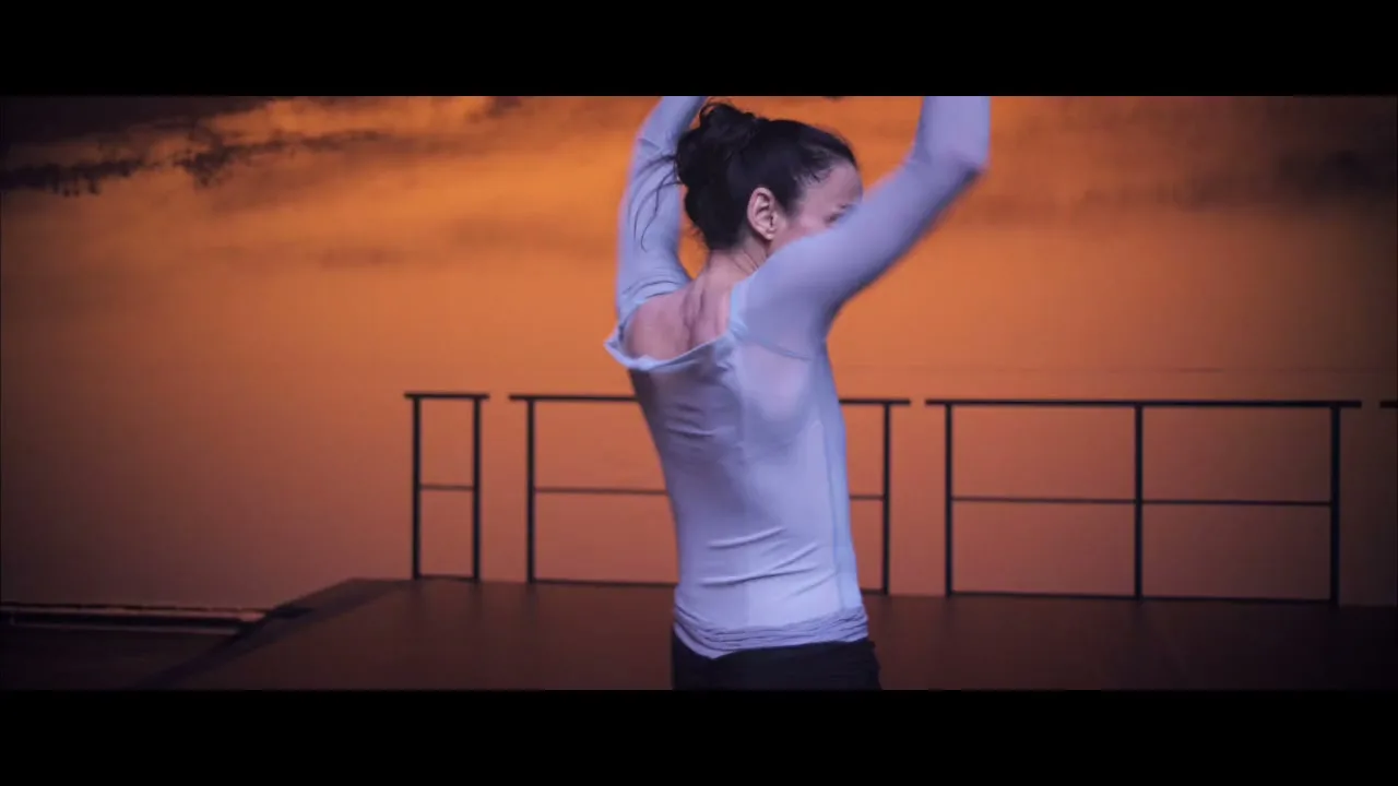 A female dancer in a light blue top and dark pants performs a ballet routine on a stage with a warm, hazy background.