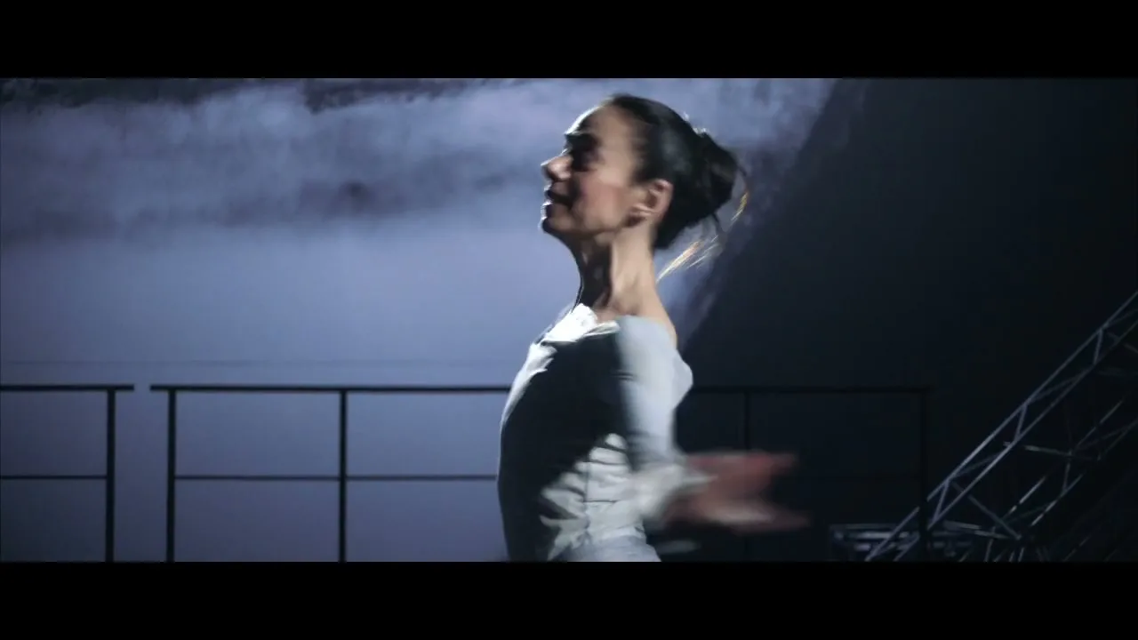 A female dancer in a grey top performs a ballet routine, turning and moving her arms gracefully in a dark, smoky studio.