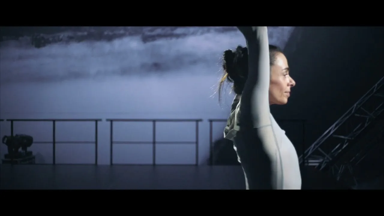 A female dancer in a grey leotard performs ballet movements on a dark stage with a projected cloudy background.