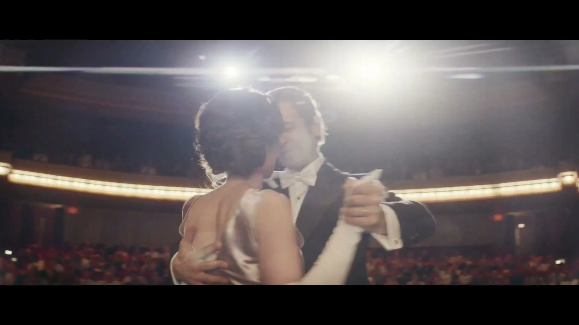 A man and woman in formal wear dance ballroom style on a brightly lit stage, with an audience visible in the background.