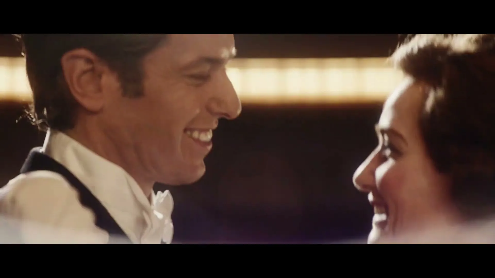A man in a tuxedo and a woman in a white dress and gloves dance closely, smiling at each other in a dimly lit room.