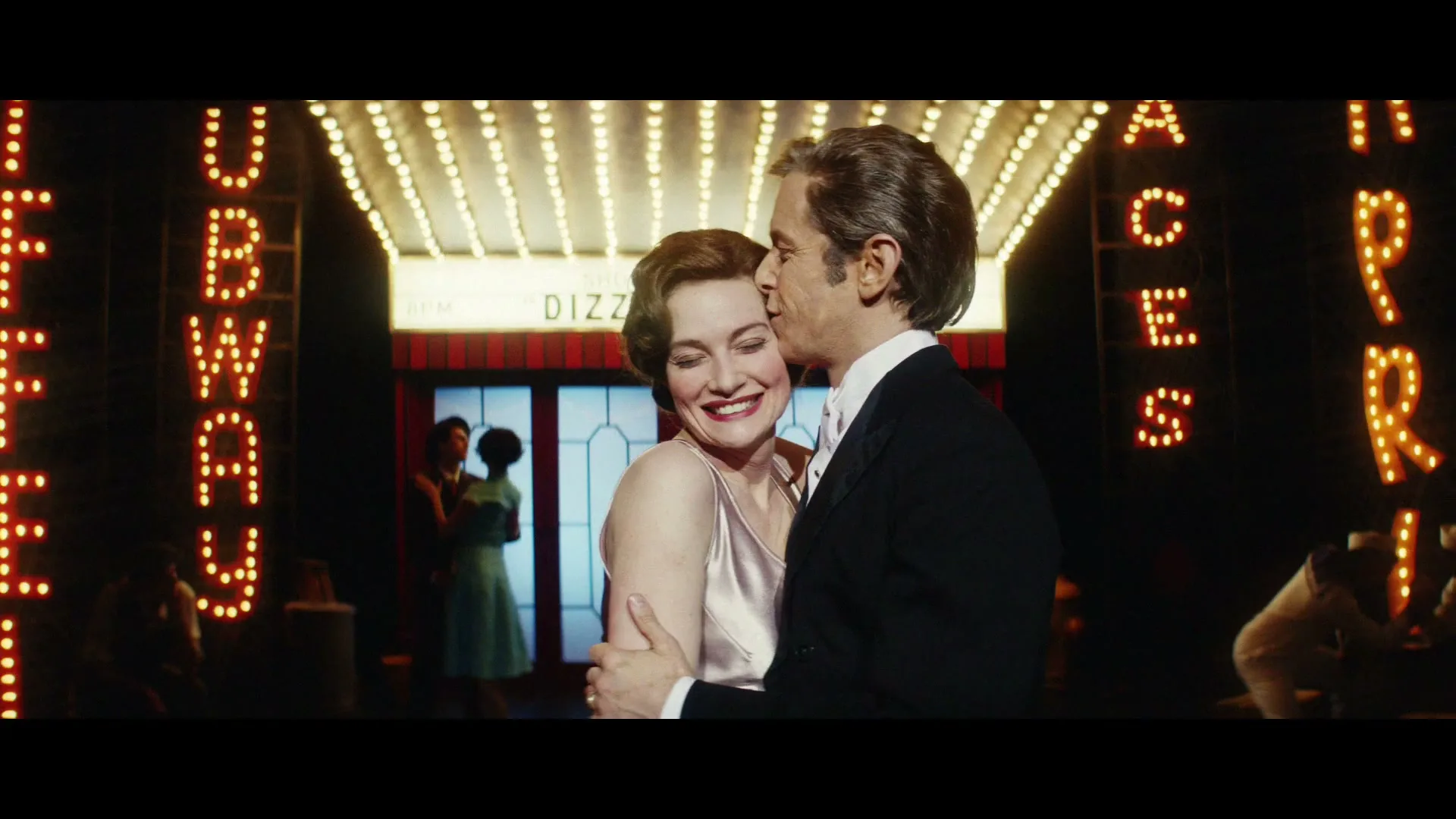 A man in a tuxedo kisses a woman in a satin dress on the cheek, both smiling, in front of a brightly lit theater marquee at night.