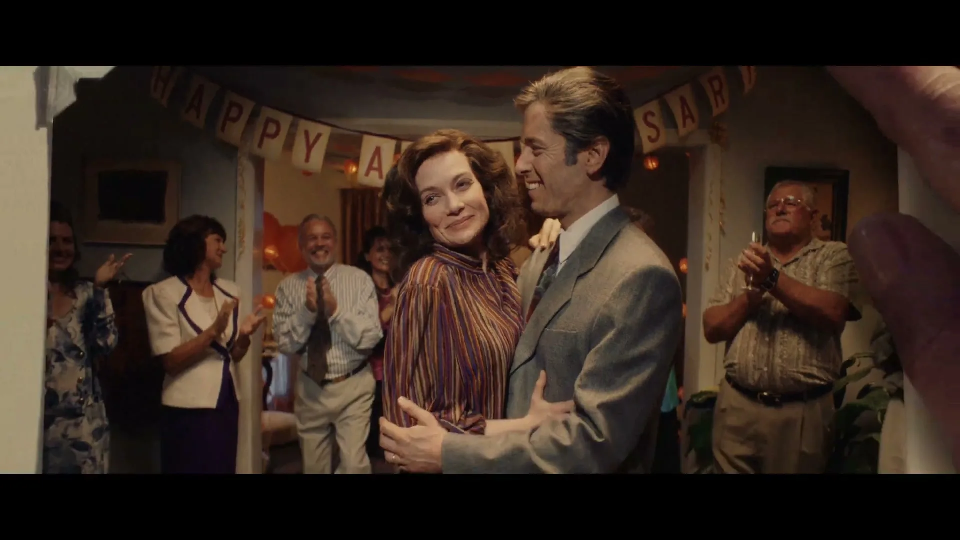 A man in a suit kisses a woman on the forehead as they embrace, surrounded by smiling guests at an anniversary party.