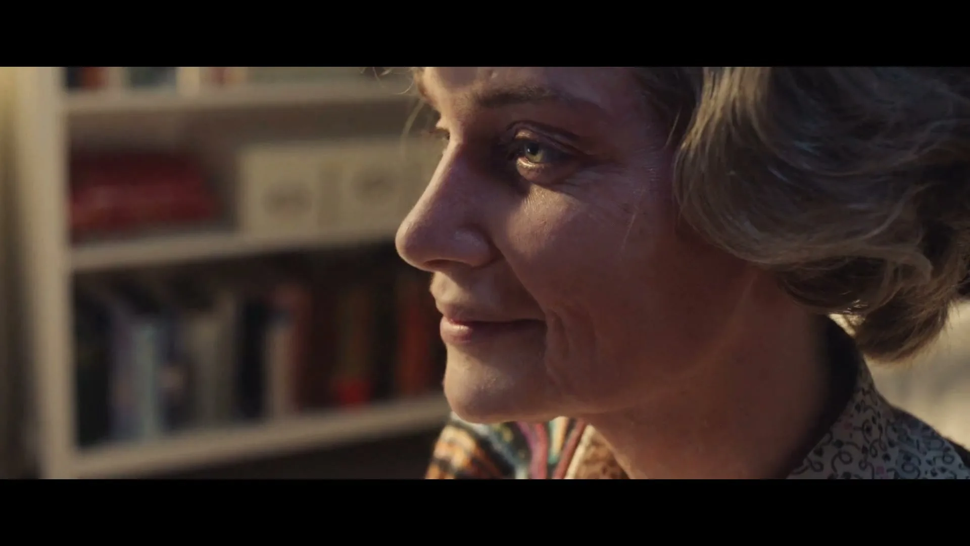 A middle-aged woman with blonde hair smiles softly, looking off-camera in a warmly lit interior with bookshelves in the background.