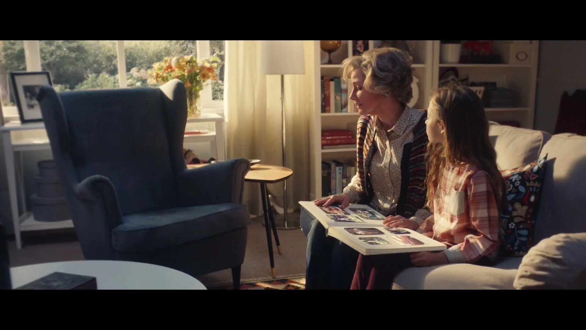 A grandmother and granddaughter sit on a couch in a cozy living room, looking at a photo album together.