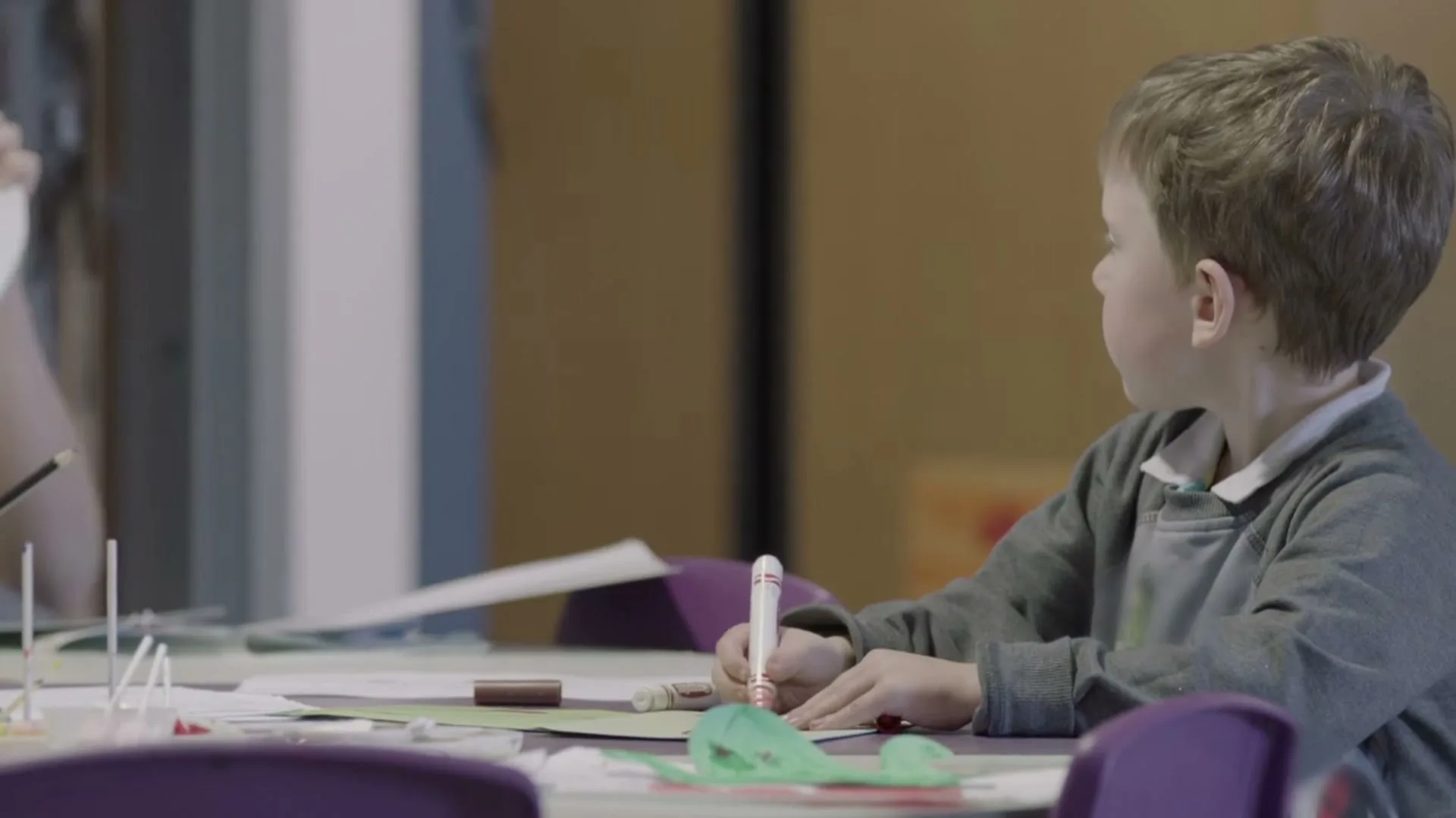 A young boy with light brown hair sits at a table, focused on drawing with a marker in a classroom setting.