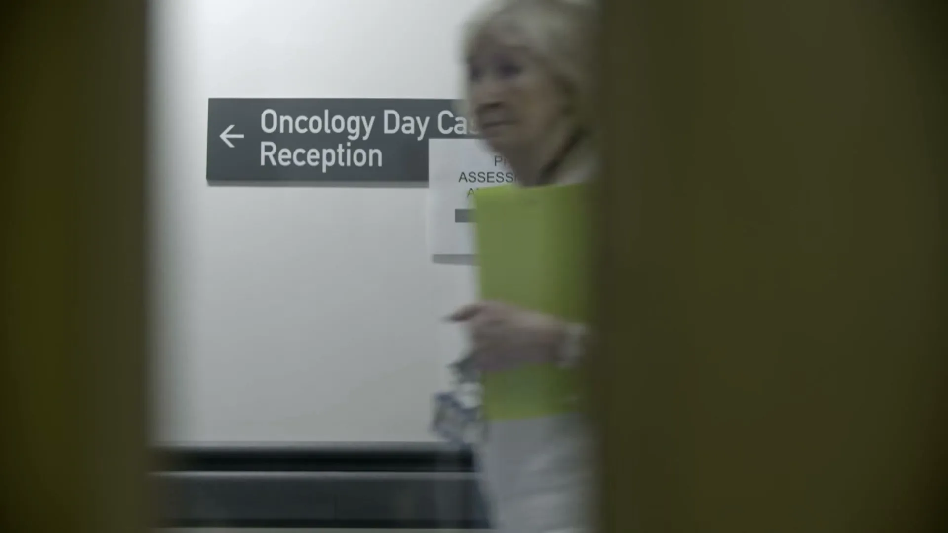 A blurred shot of a hospital corridor with a sign for "Oncology Day Case Unit Reception" and a woman walking past, seen through a doorway.