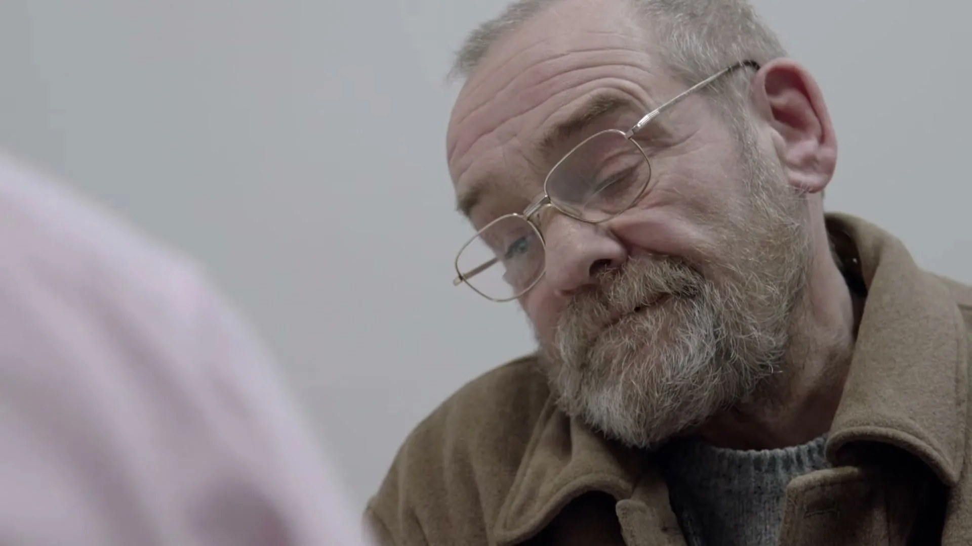 A man with a beard and glasses looks up at someone off-screen, listening intently in an interior setting.