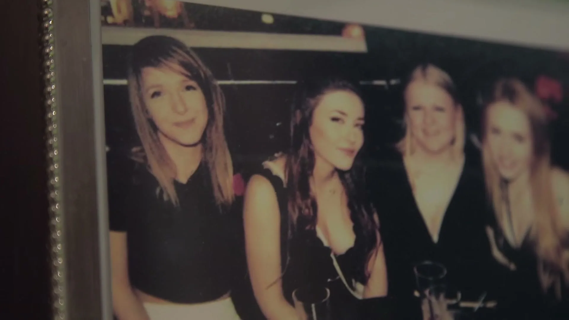 A close-up shot of a framed photograph shows four young women smiling at a party, with one woman in the foreground looking directly at the camera.