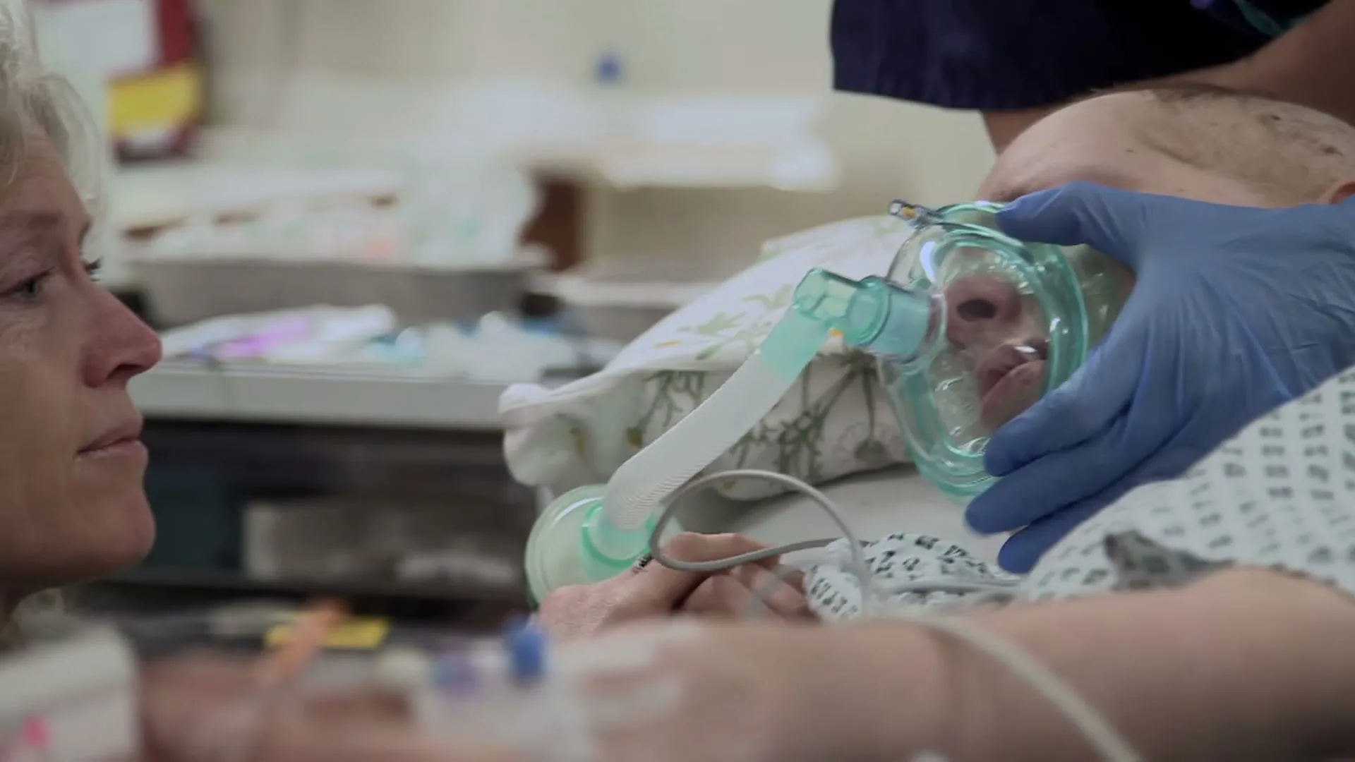 A nurse in blue gloves adjusts an oxygen mask on a patient in a hospital bed, while a woman with curly grey hair watches intently.