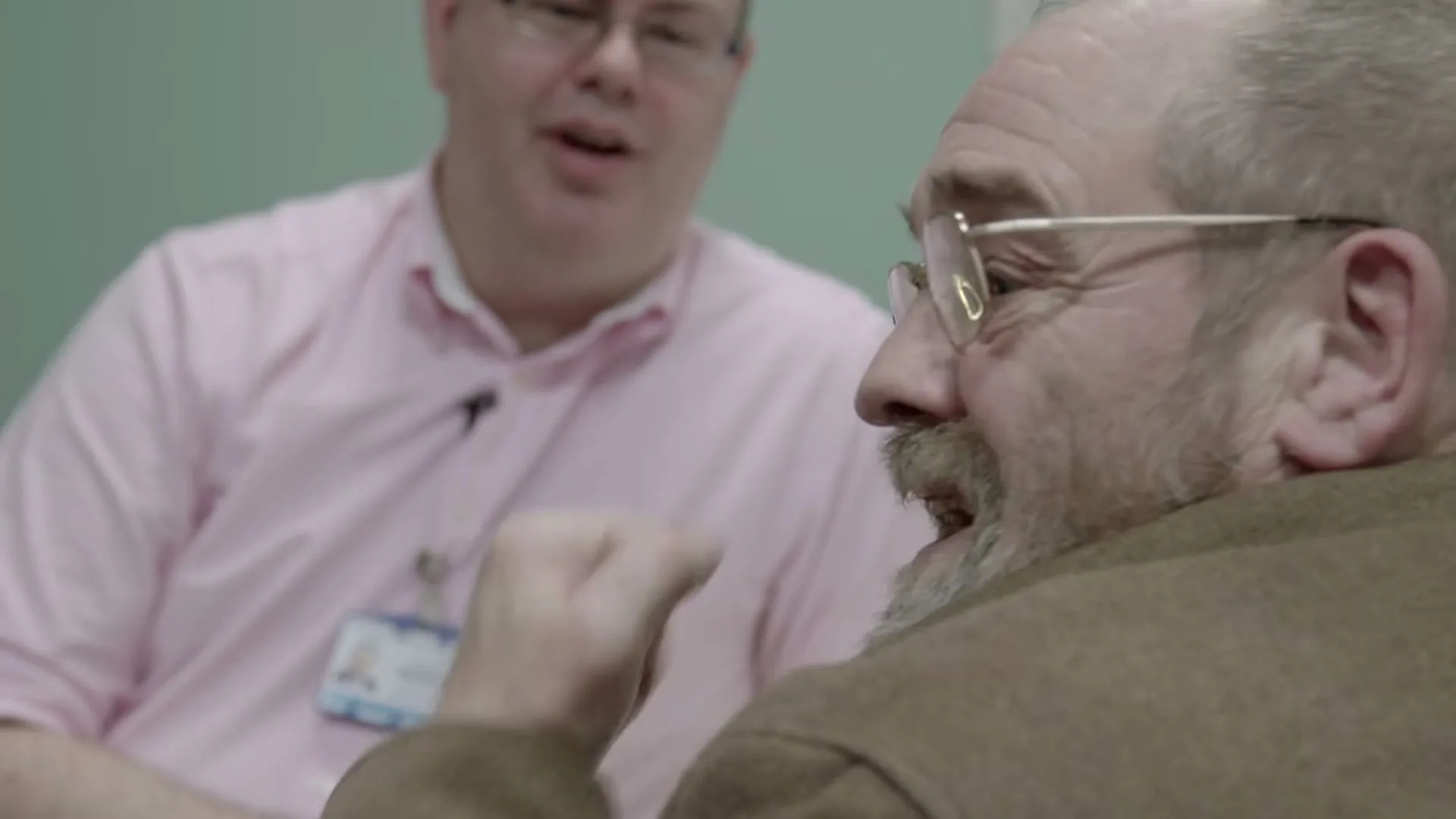 A male patient with glasses and a beard talks to a male doctor in a pink shirt, both smiling in a hospital room.