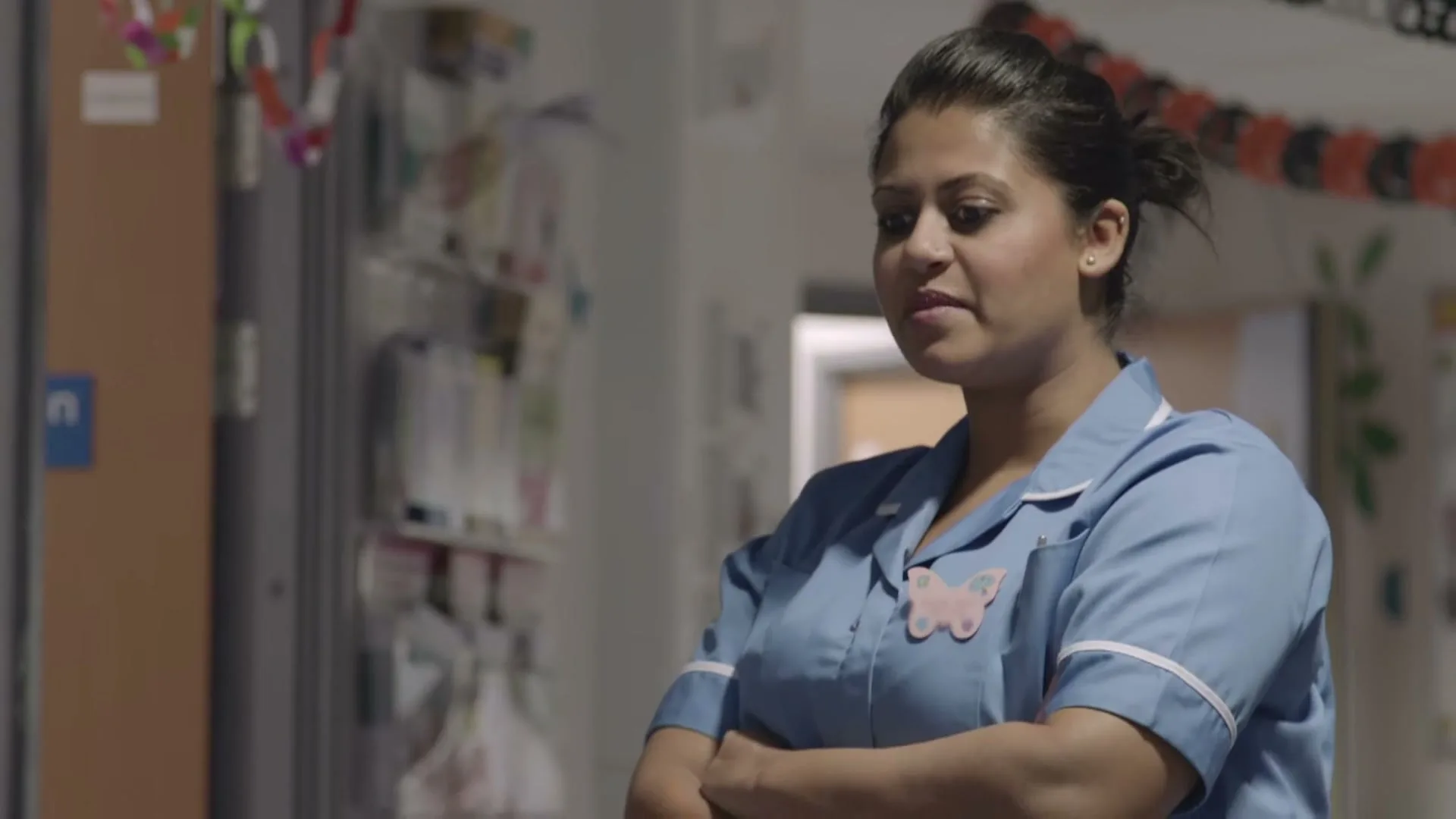 A female nurse in blue scrubs with a butterfly pin smiles and crosses her arms in a hospital hallway decorated with paper chains.