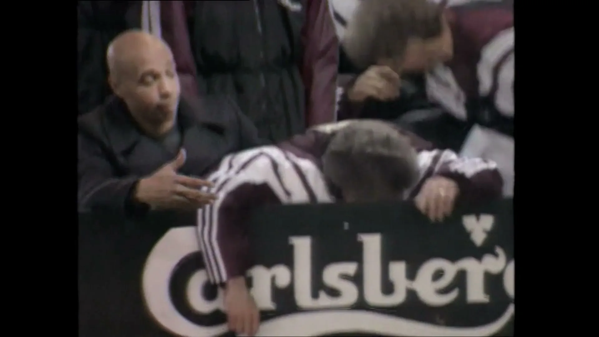 A man in a maroon and white tracksuit leans over a "Carlsberg" sign, looking distraught, while another man in a dark coat looks on at a football match.