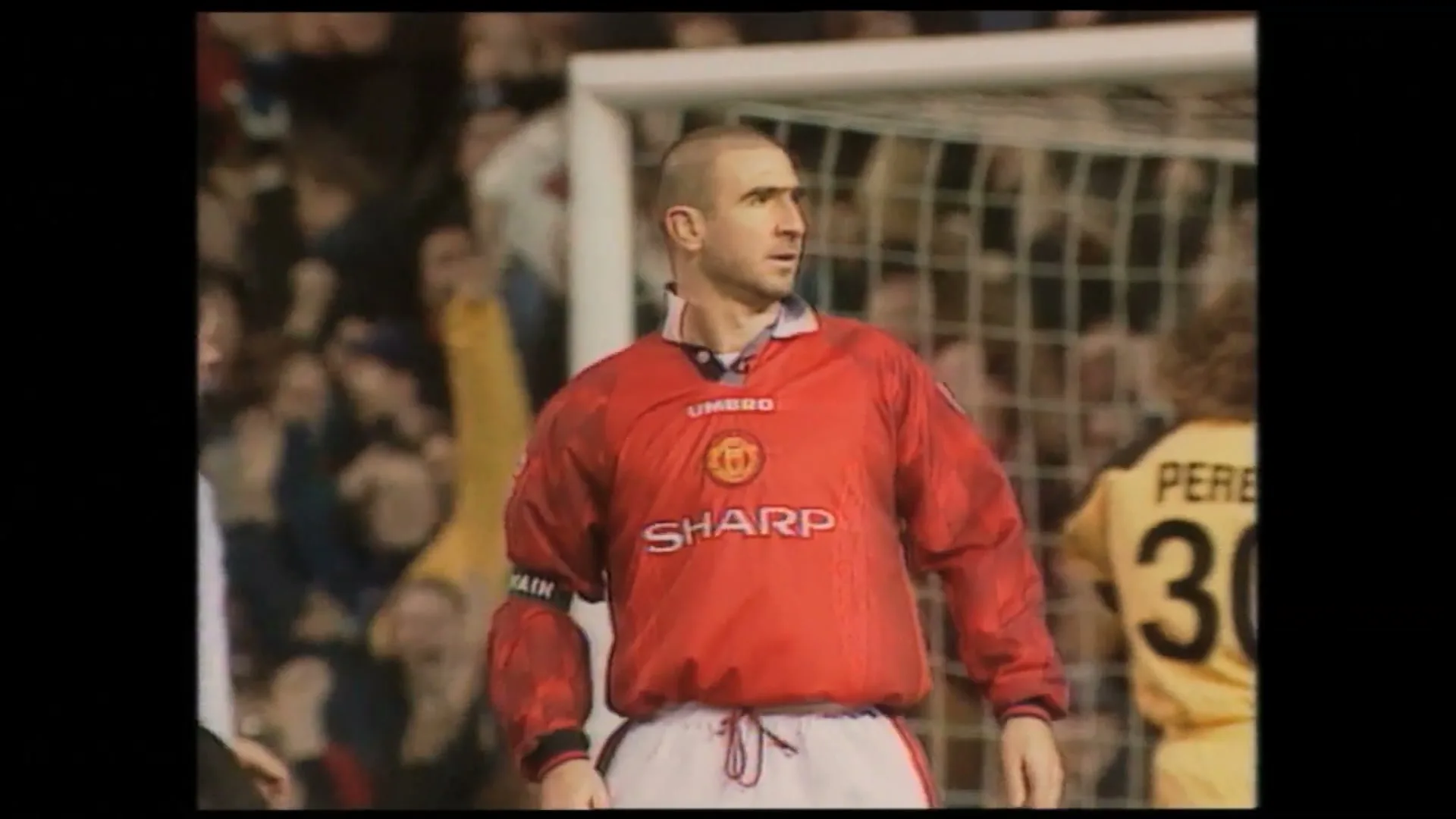 A bald male soccer player in a red jersey stands on a field, looking around during a game in a stadium.