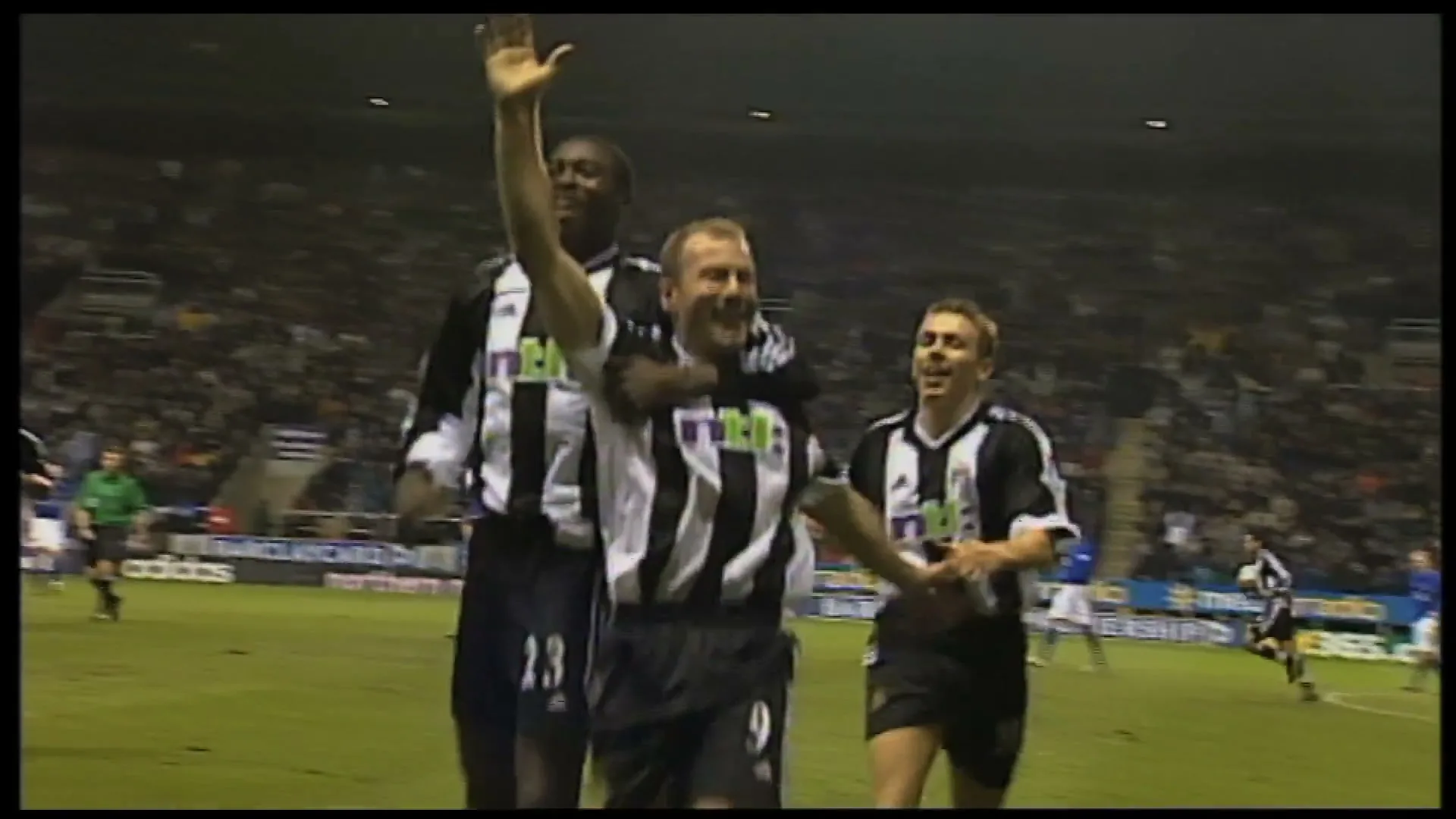 Three male soccer players in black and white striped jerseys celebrate a goal on a green field in a stadium at night.