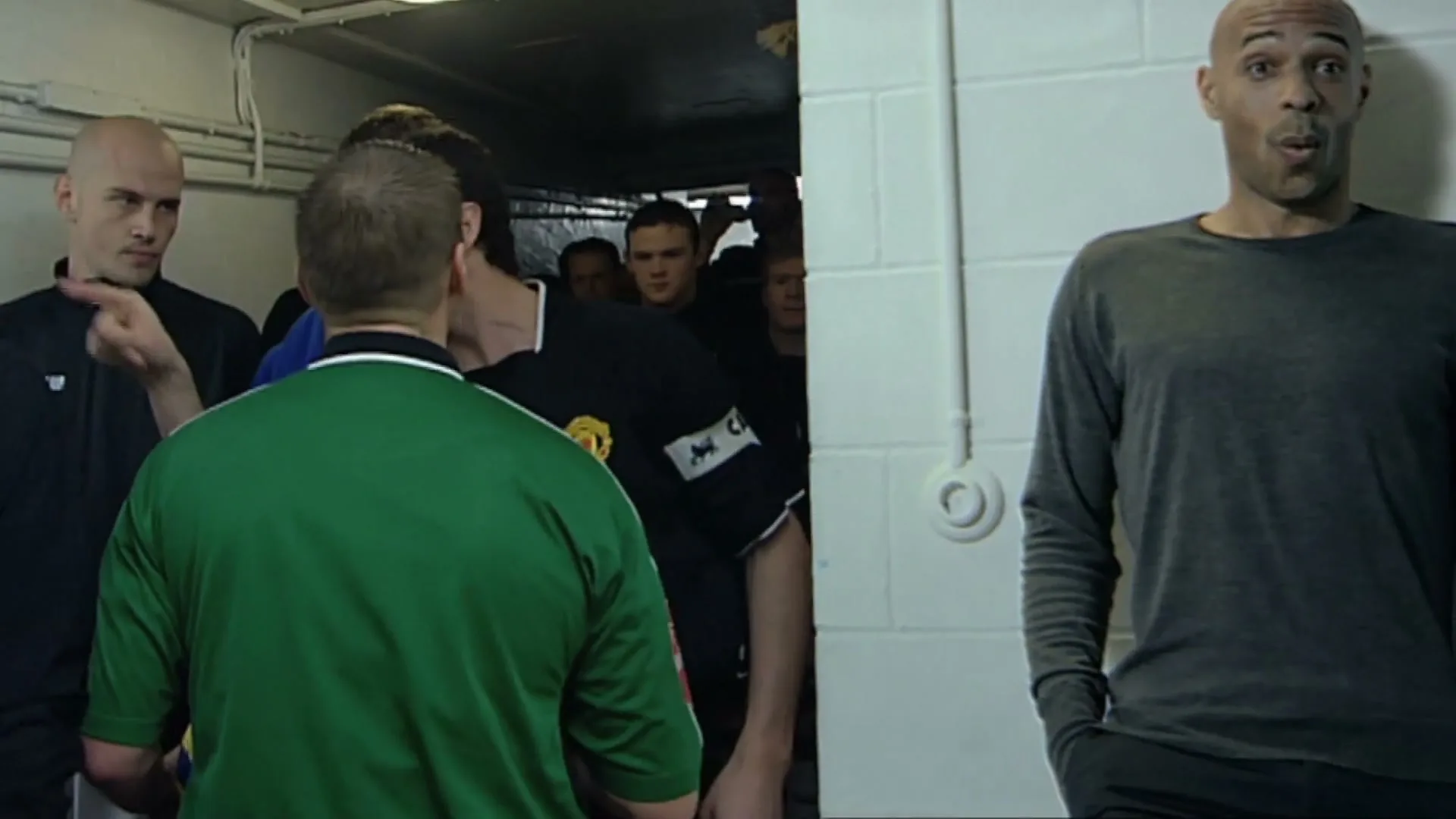 Roy Keane, wearing a black Manchester United jersey, argues with a referee in a green jersey in a stadium tunnel, while Thierry Henry watches from the side.