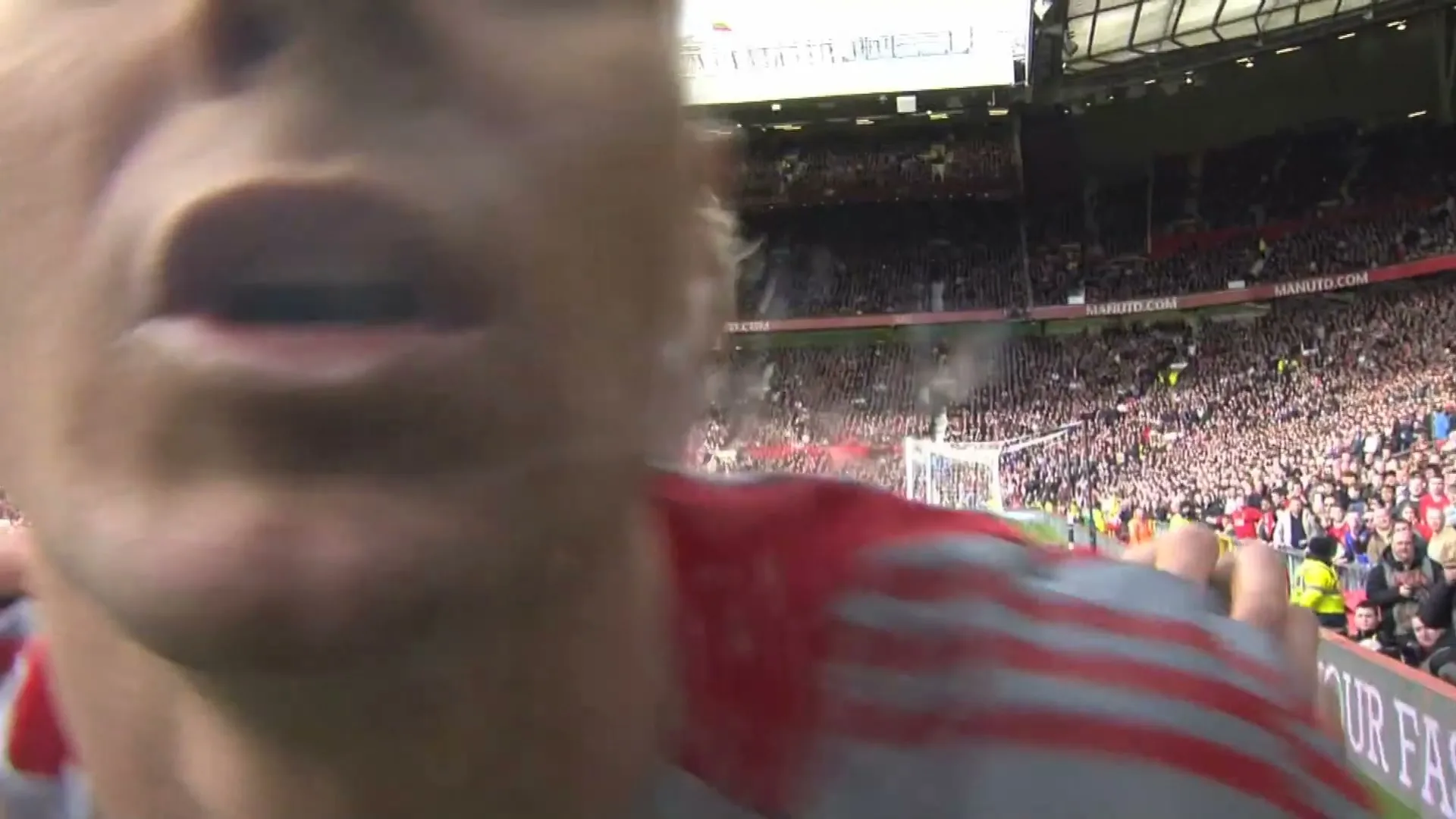A male football player in a red and white jersey celebrates a goal, shouting into a camera held very close to his face on a sunny day in a stadium.