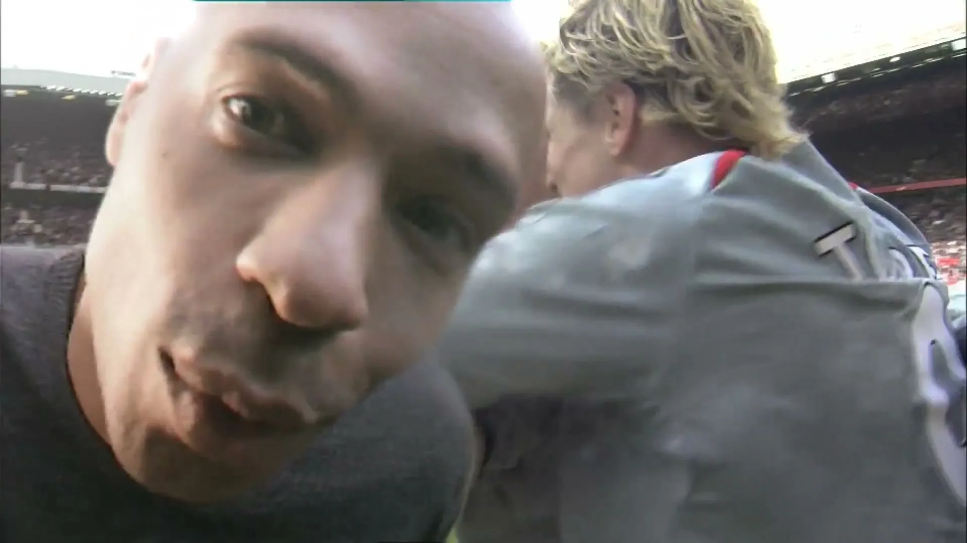 A bald man in a dark t-shirt holds a camera up close to his face, capturing a group of male soccer players in grey jerseys celebrating behind him on a sunny day in a stadium.