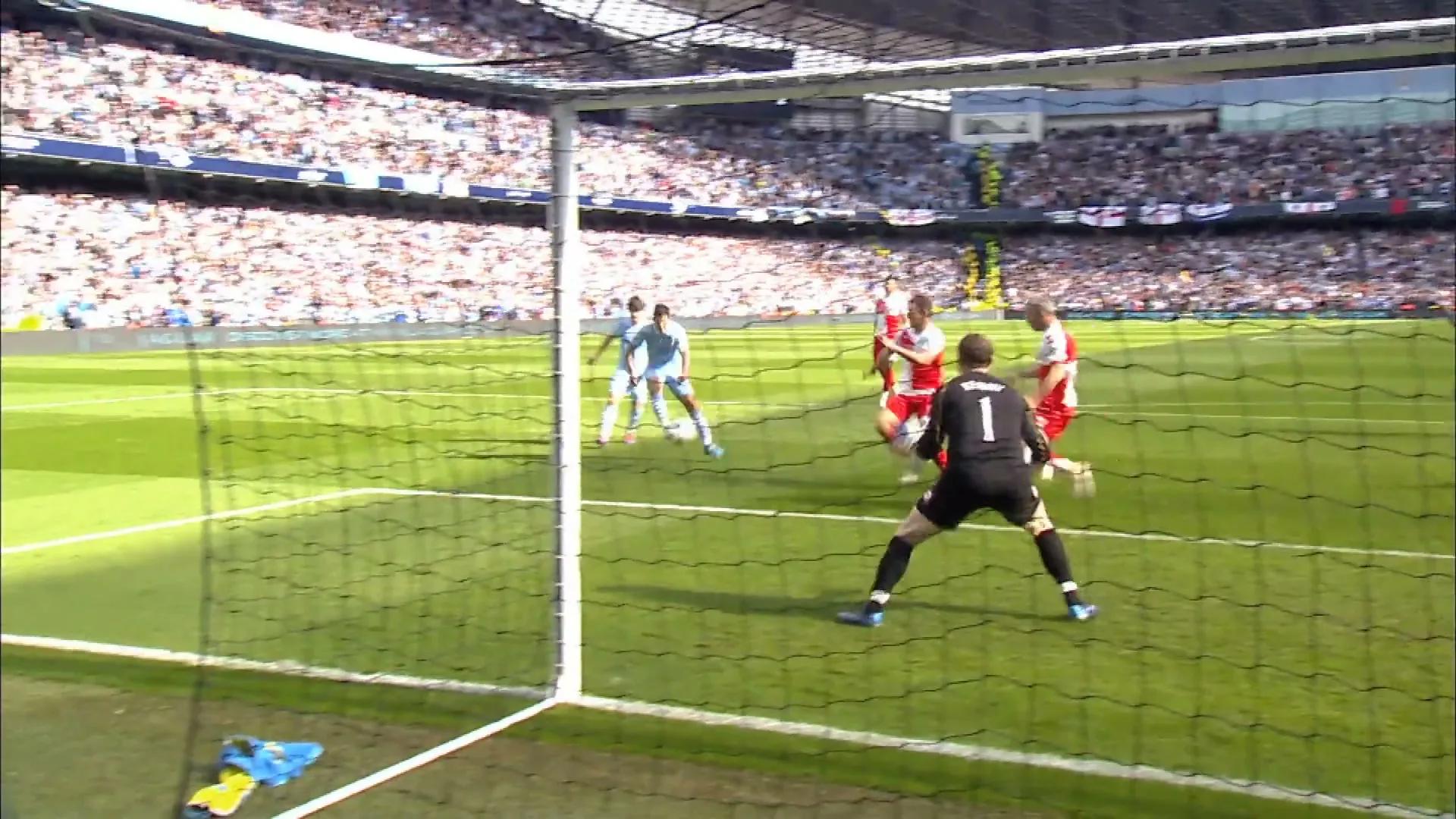 A wide shot from behind the goal shows a soccer game in progress, with players in light blue and red uniforms on a green field, and a large crowd in the stands.