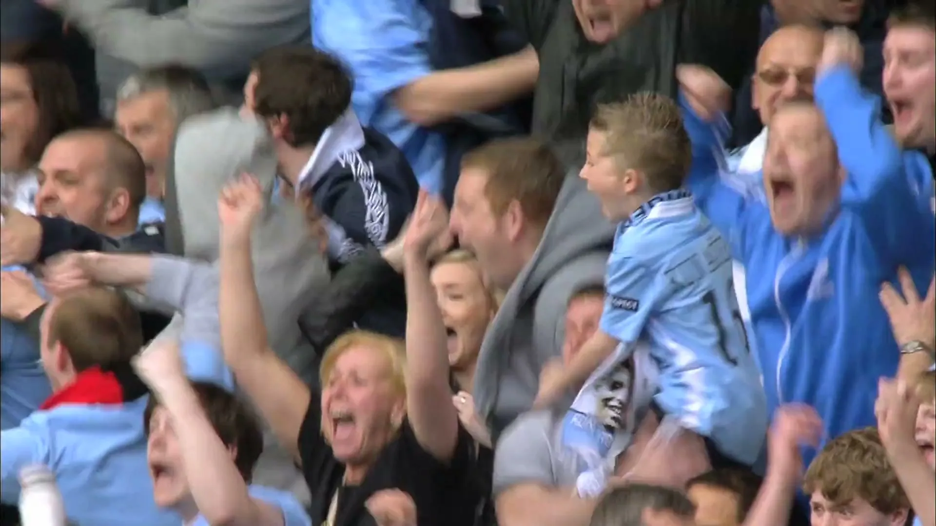 A crowd of excited football fans, wearing light blue jerseys, cheer and raise their arms in a stadium.