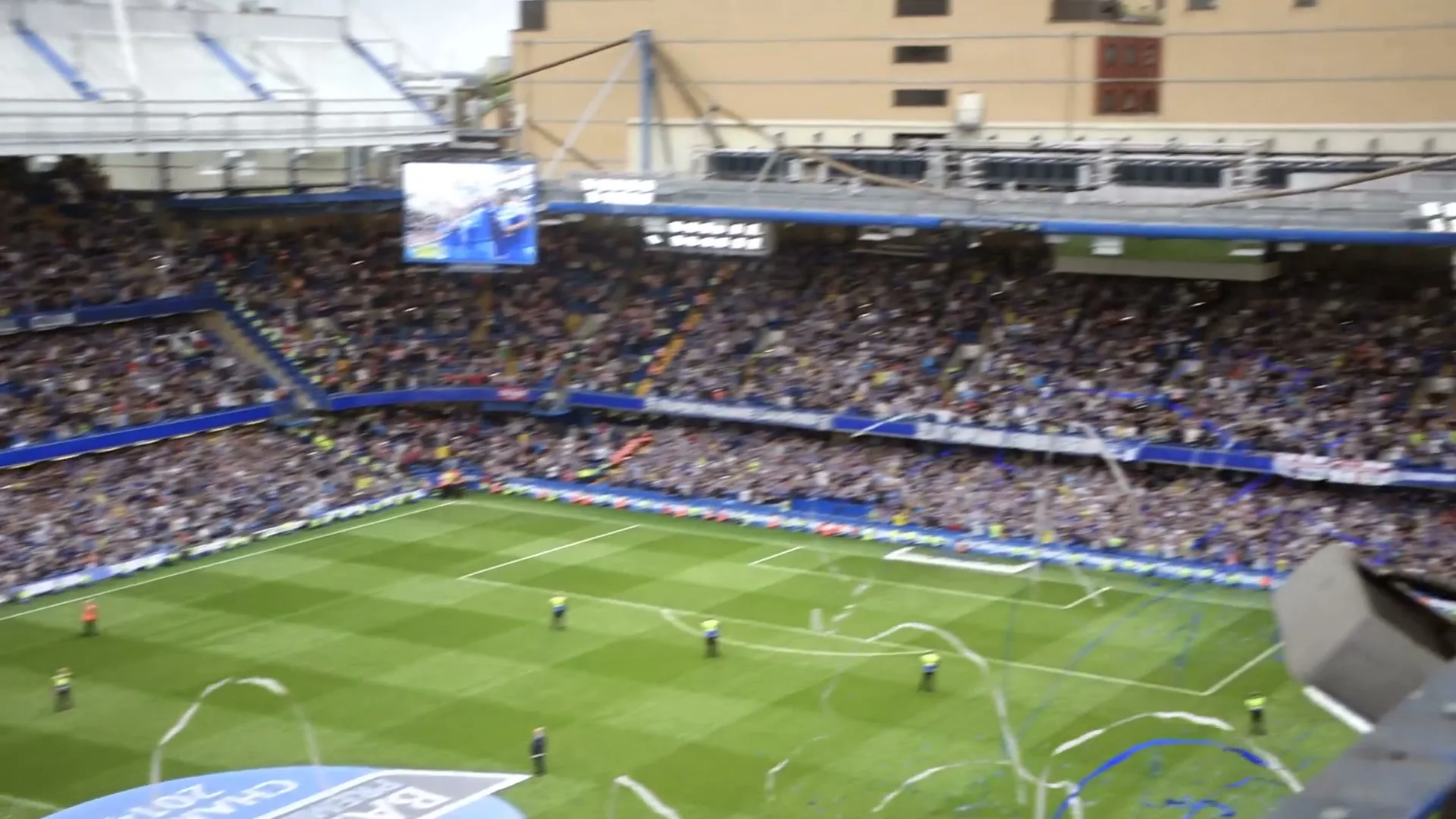 Thierry Henry, a bald man in his 30s, wearing a black long-sleeved shirt, smiles at the camera from a high vantage point overlooking a crowded football stadium during a celebration.