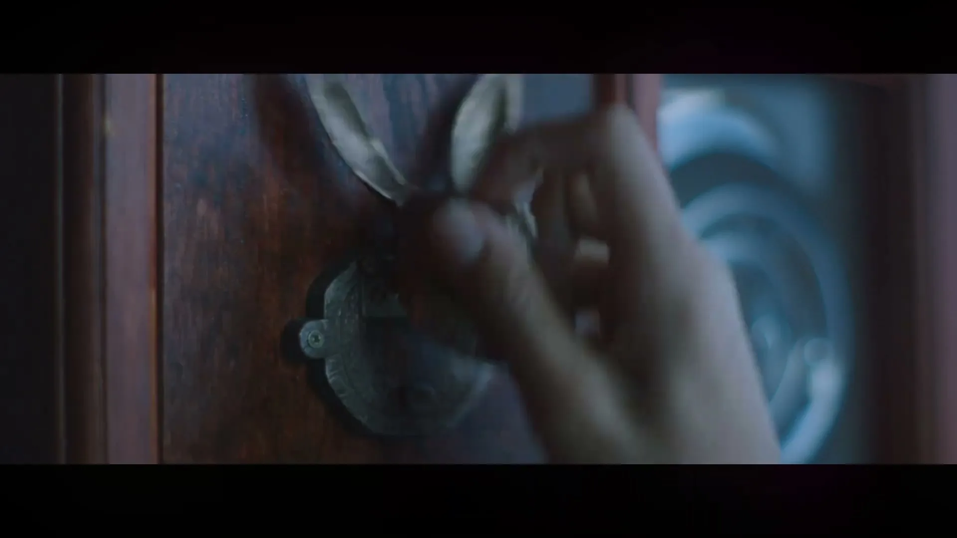 A child's hand reaches out to touch a dark, metallic hare head door knocker on a wooden door in a dimly lit interior.
