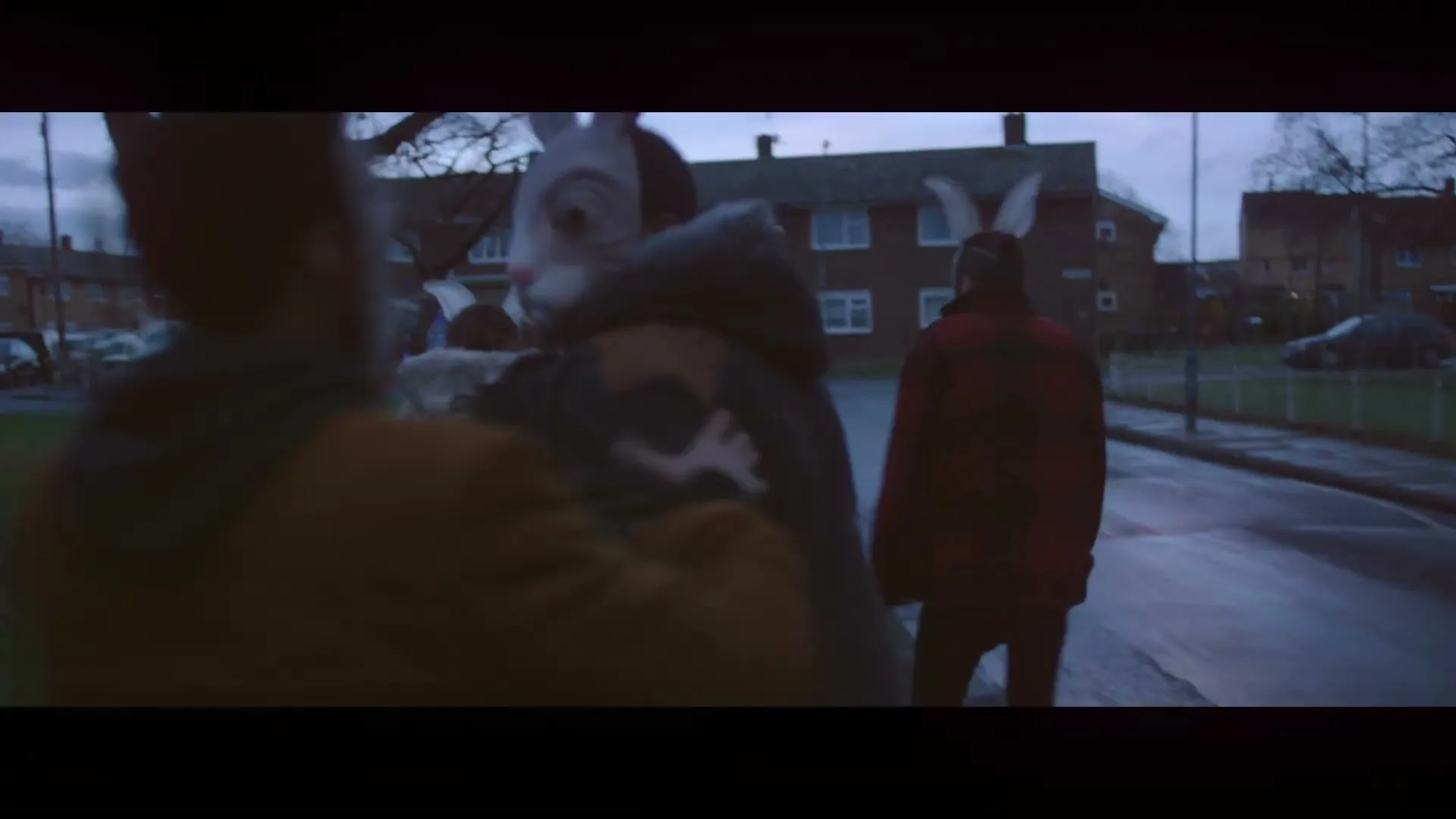 Three people in rabbit masks walk away from the camera down a suburban street at dusk.