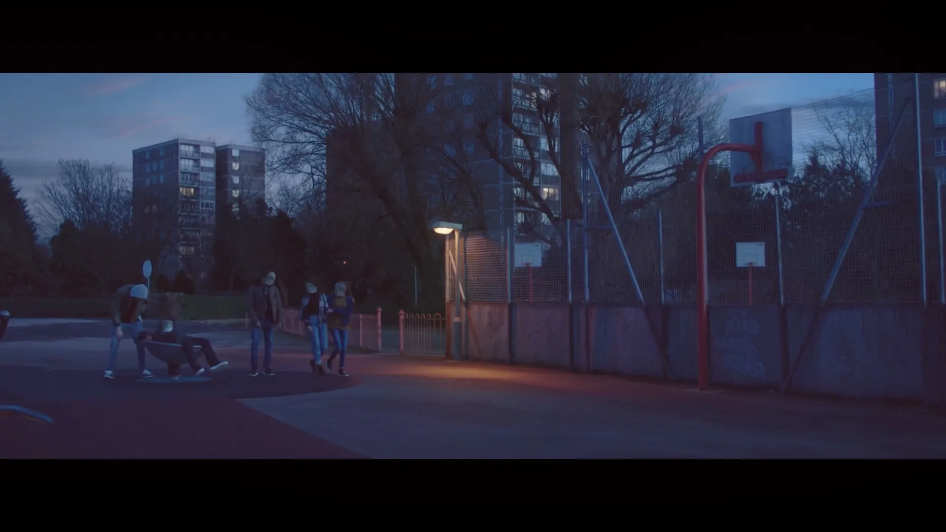 A group of teenagers walk past a boy sitting in a wheelchair on a basketball court at dusk, with apartment buildings in the background.