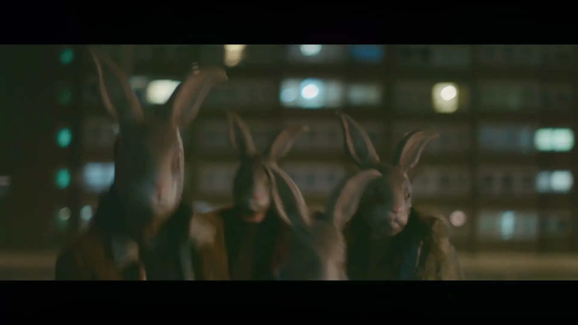 A group of people wearing rabbit masks and casual clothes run towards the camera at night, with blurred city lights in the background.
