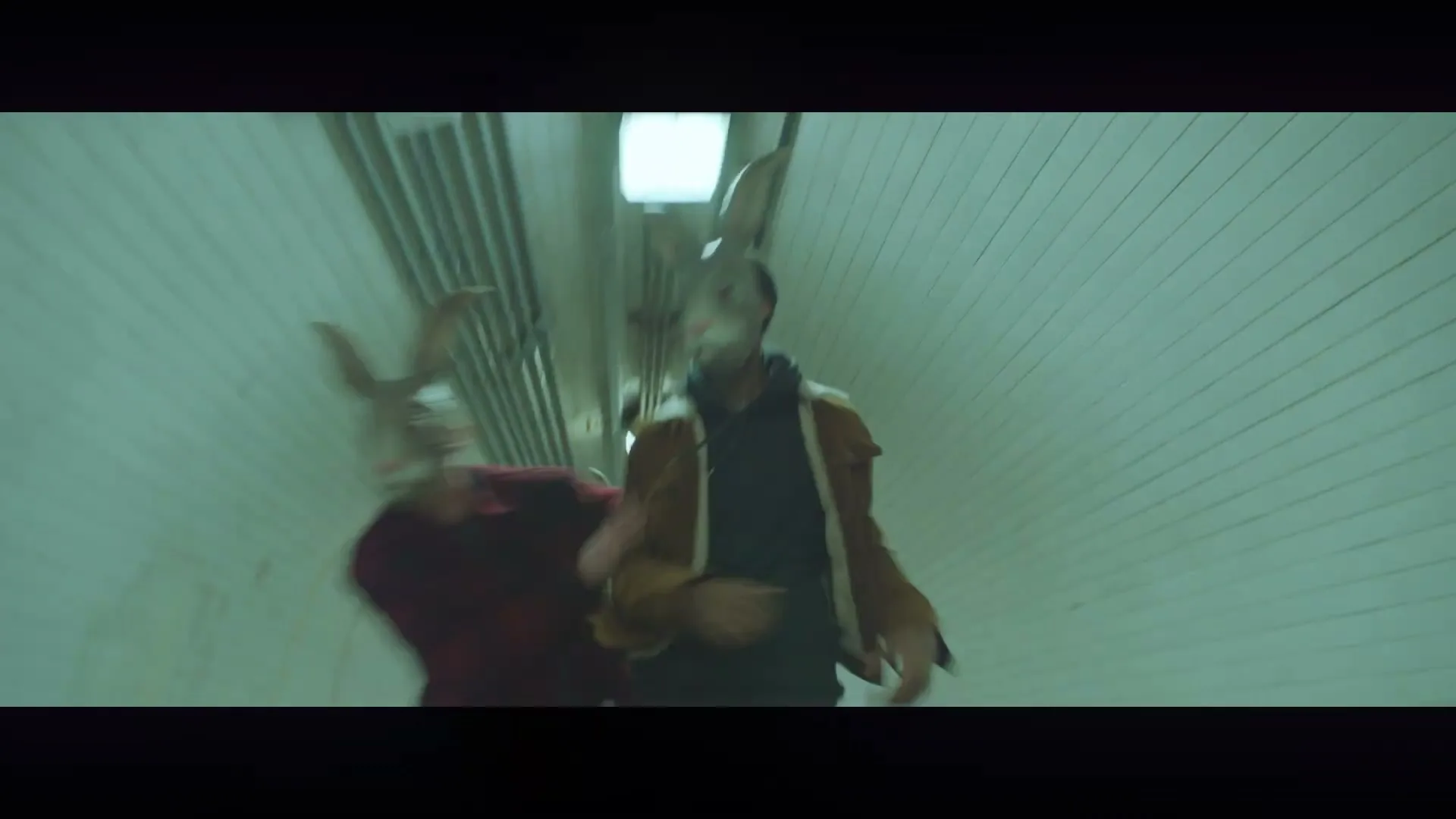 Two people in rabbit masks run through a green-tiled underground tunnel, one in a brown jacket and the other in a red plaid shirt.