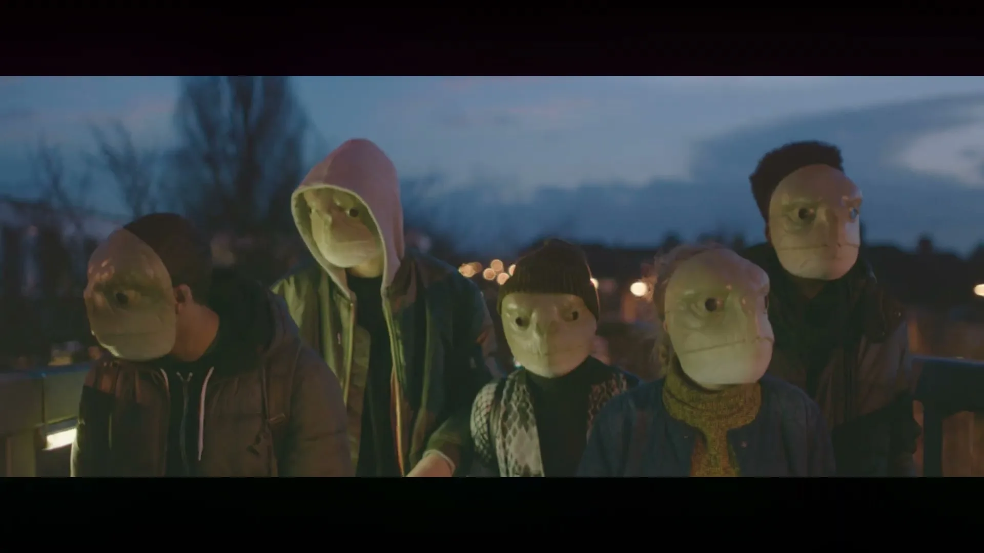 A small group of people wearing turtle masks stand on a bridge at dusk, looking down at something off-screen.