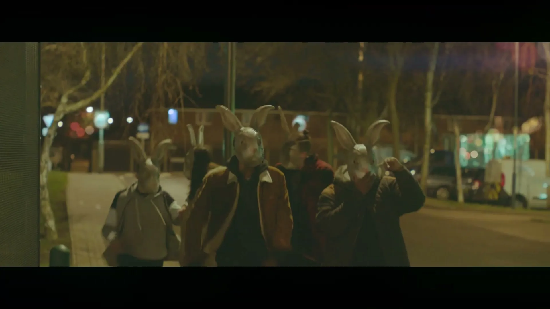 A group of young people wearing rabbit masks walk quickly down a street at night, with streetlights and buildings in the background.