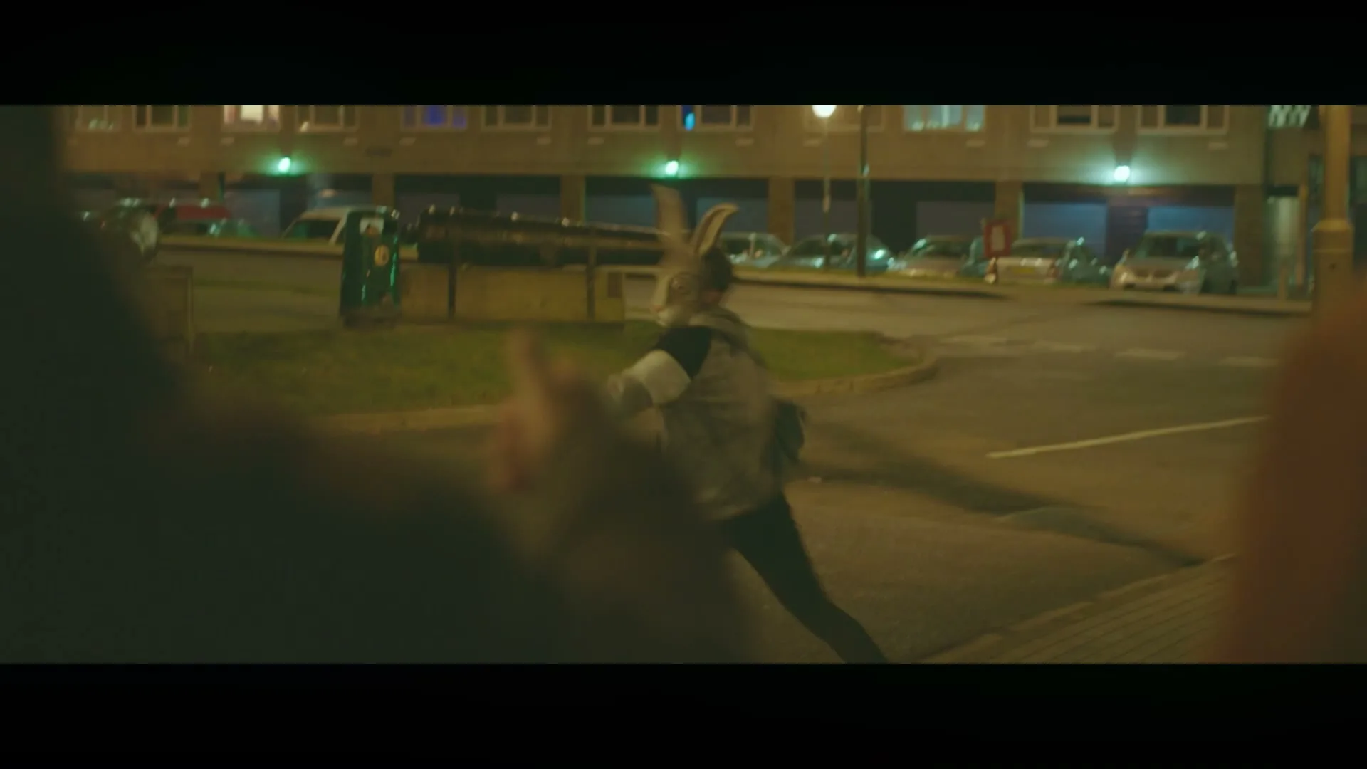 A person in a grey hoodie with rabbit ears runs across a street at night, with apartment buildings and parked cars in the background.