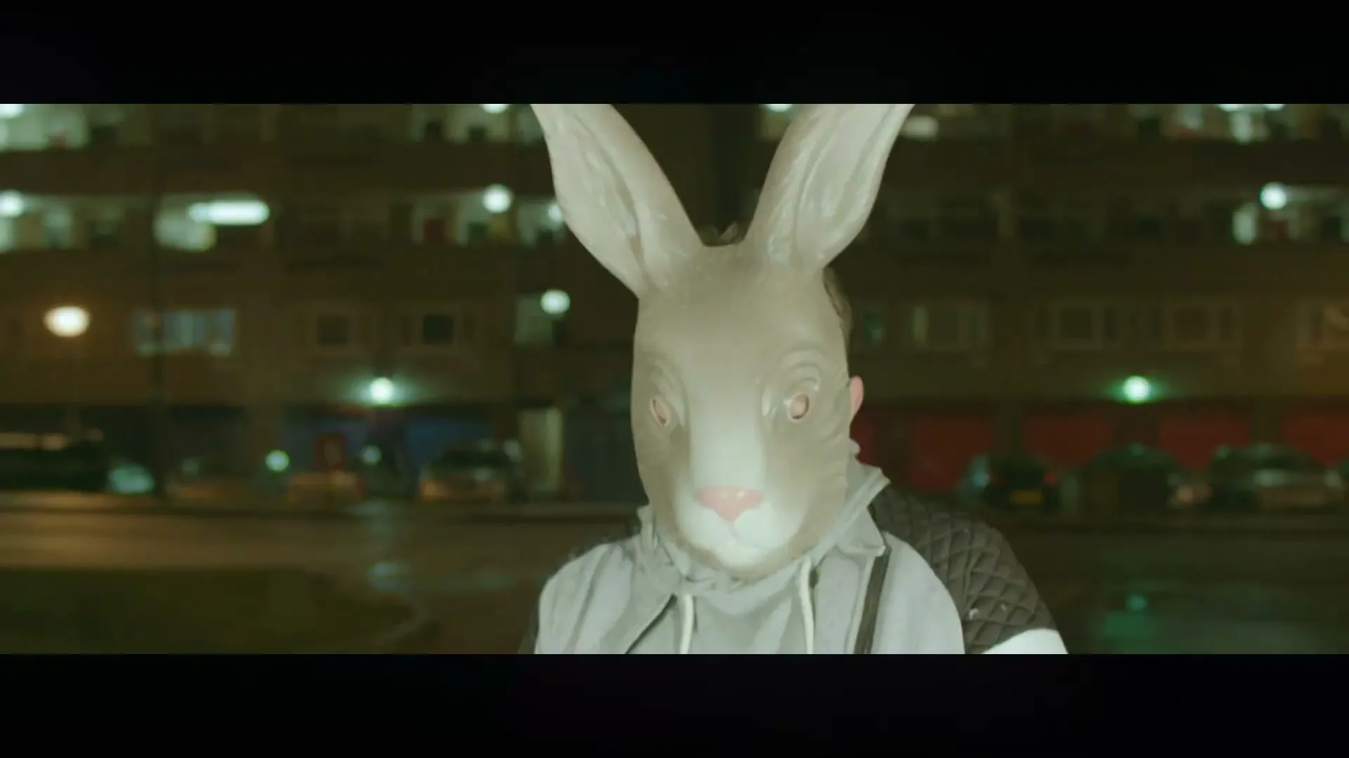 A person in a grey rabbit mask and hoodie stands still at night in front of a block of flats.