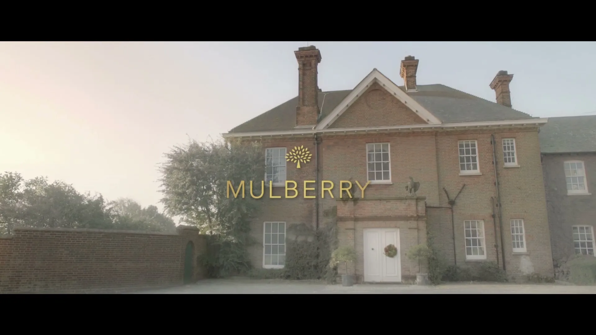 A wide shot of a large, traditional brick house with a white front door and a Christmas wreath, with the Mulberry logo superimposed on the facade.