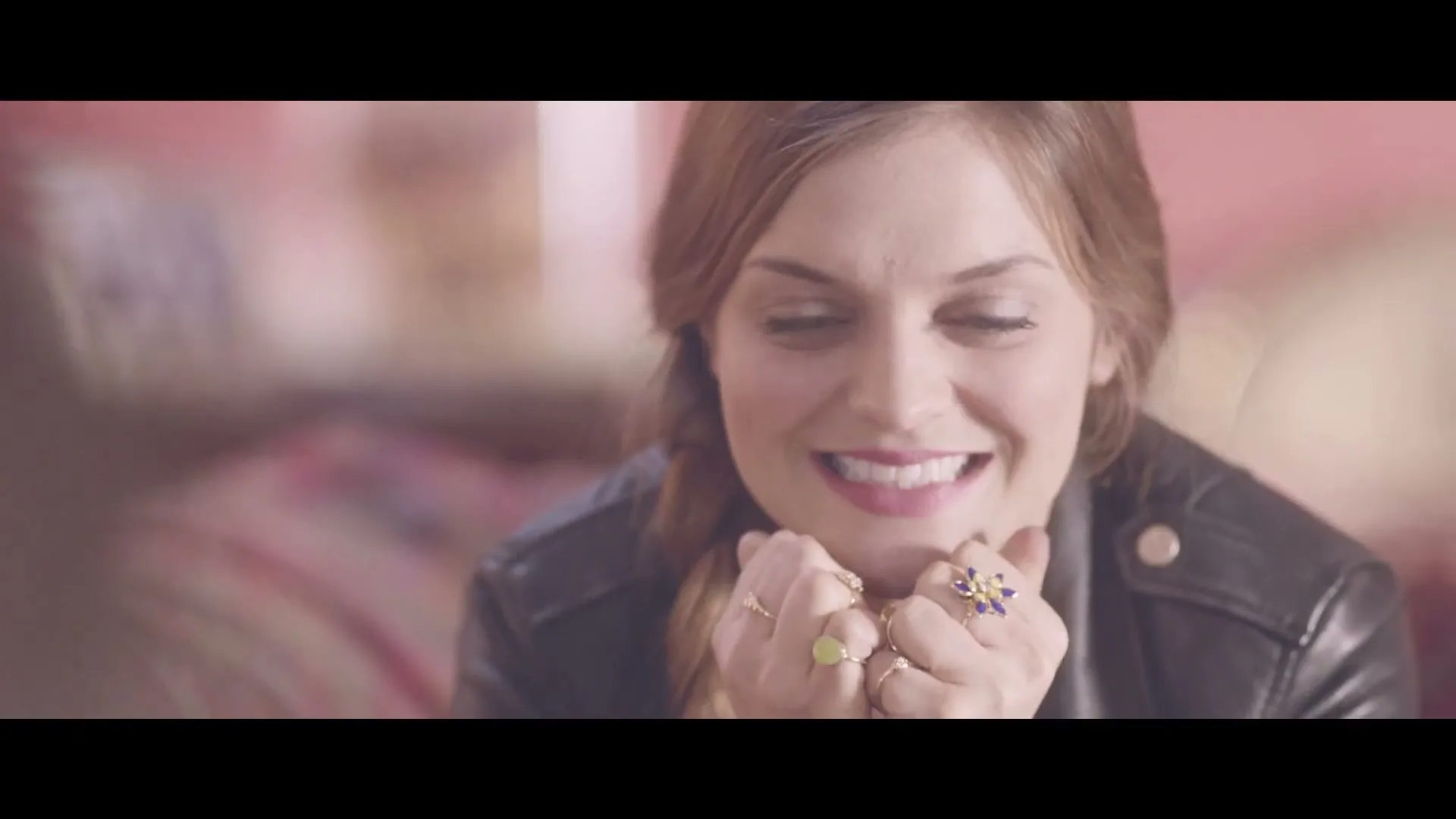 A young woman with a braid and a black leather jacket smiles, holding her hands with rings under her chin, in a soft-focus interior.