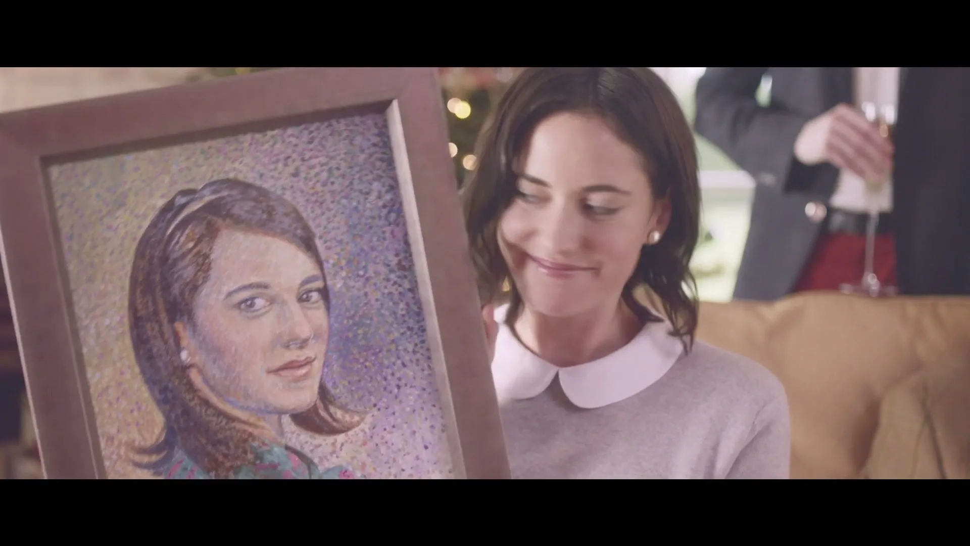 A woman with dark hair and a white collar sweater holds up a framed painting of a woman, smirking at the camera in a living room.