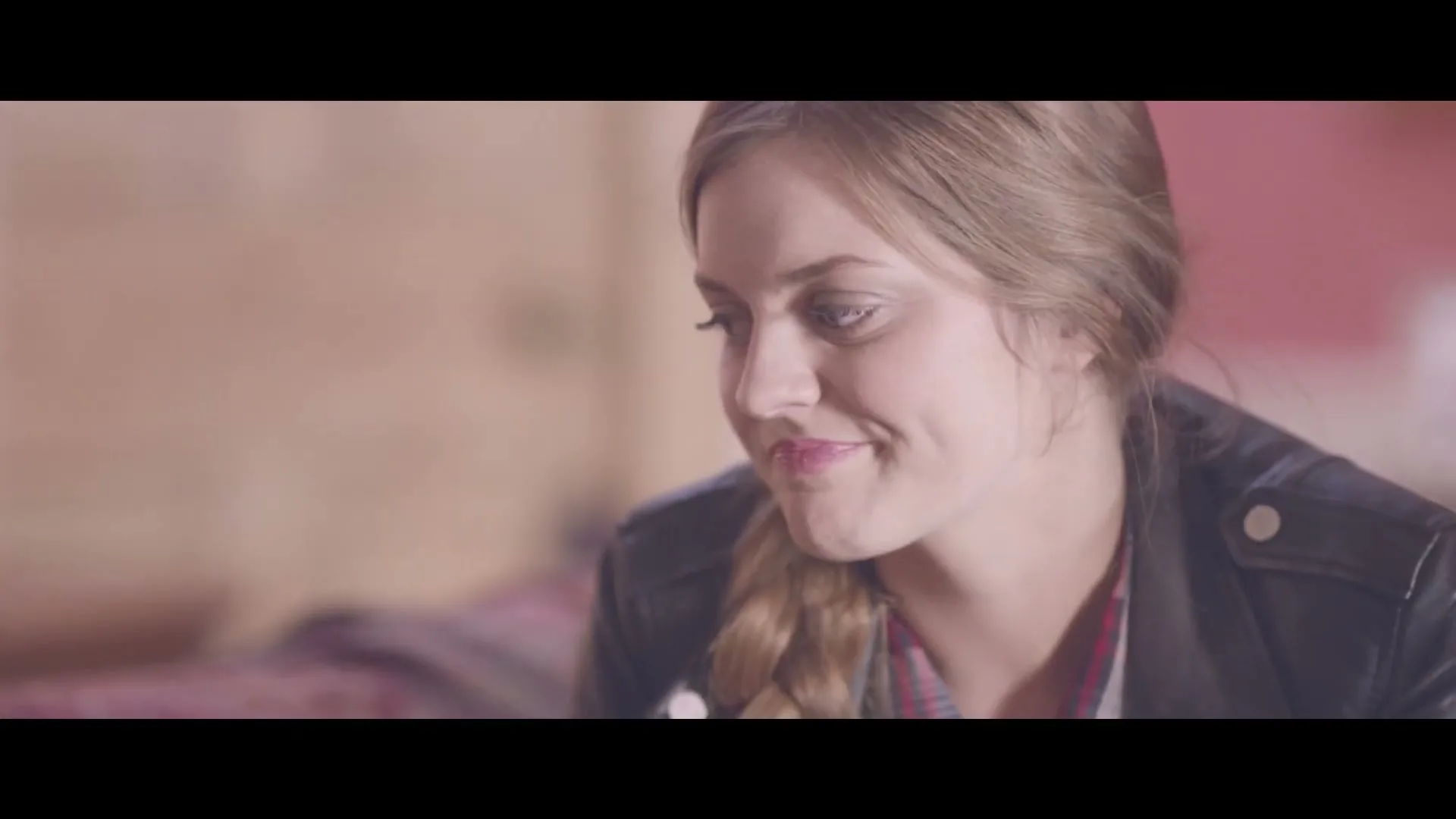 A young woman with a braid and a leather jacket smiles shyly, looking down and to the side in a softly lit interior.