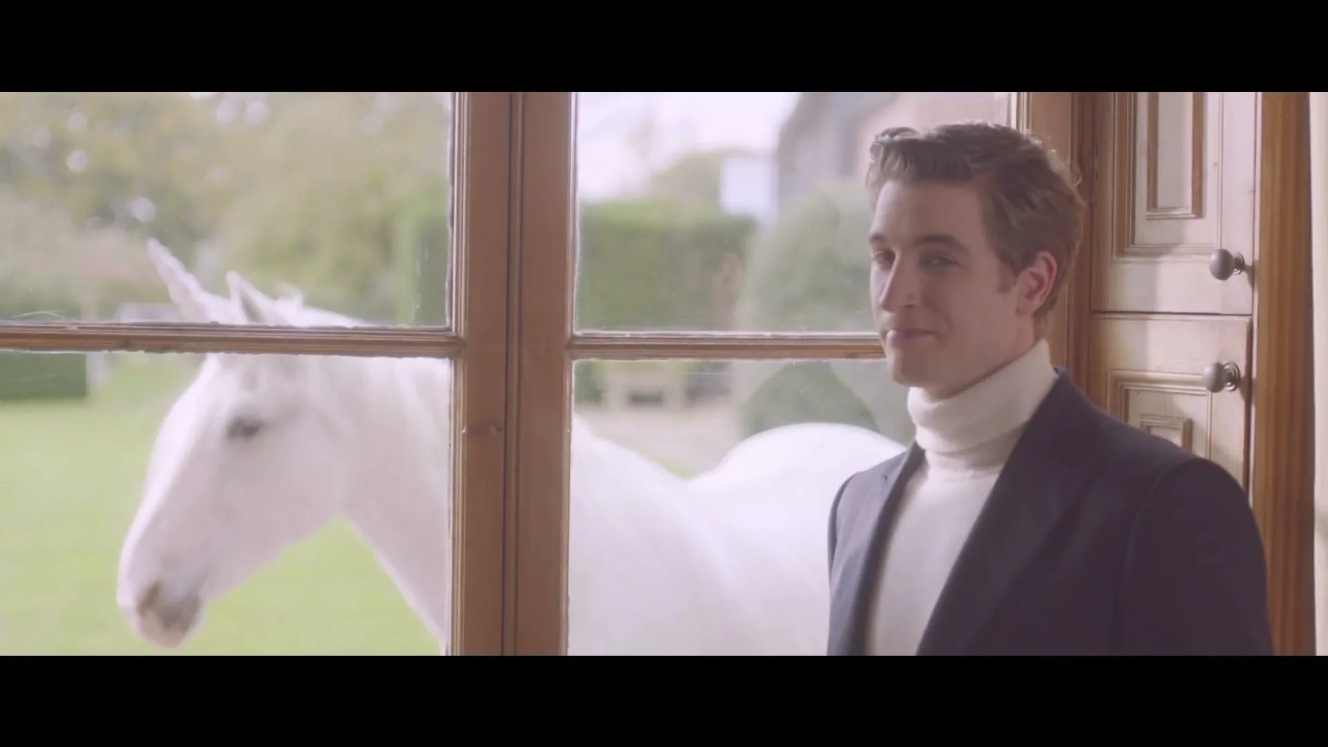 A young man in a suit and turtleneck stands by a window, looking at a white unicorn outside in a garden.