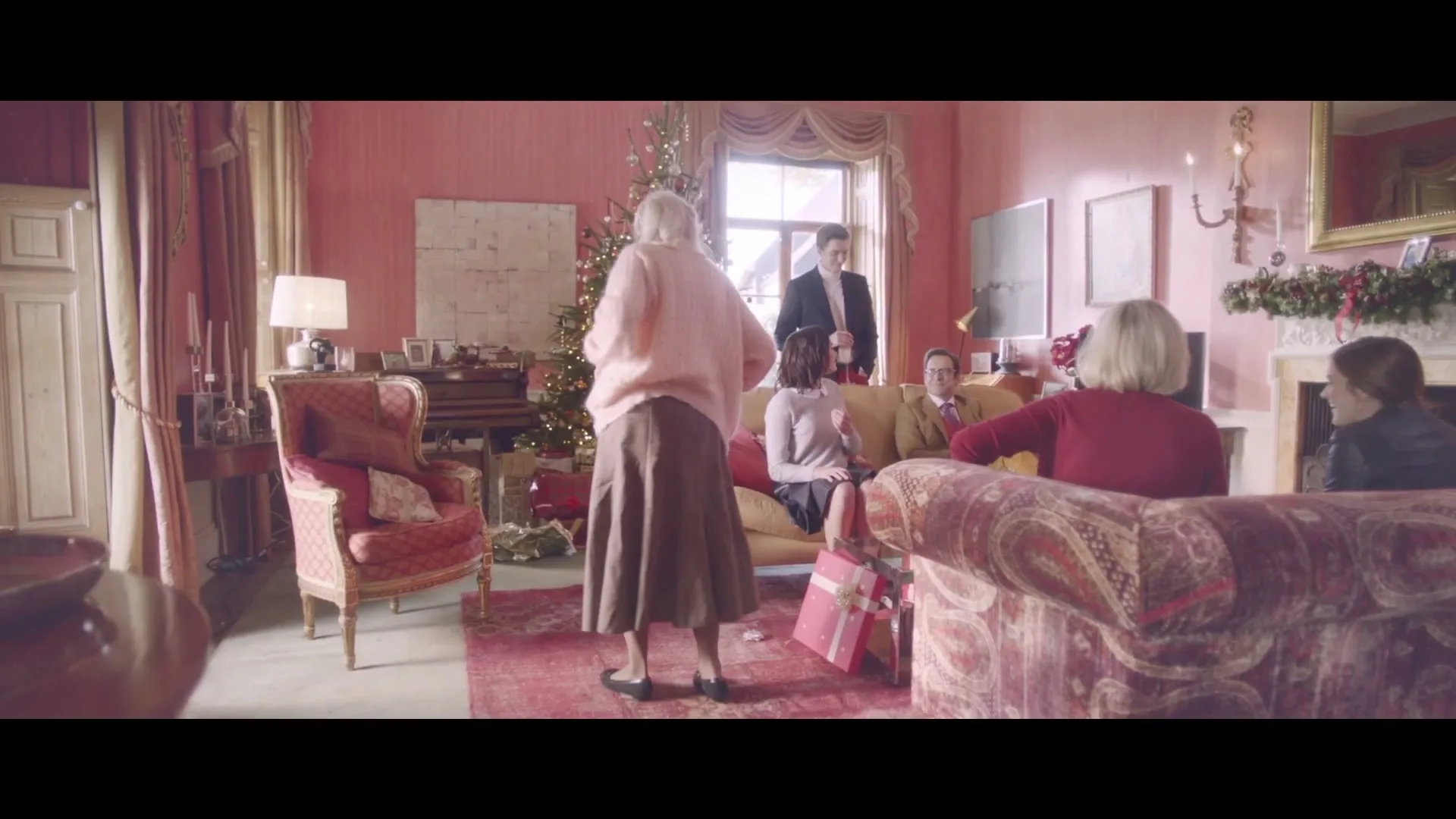 An older woman in a pink sweater and brown skirt stands in a festive living room, surrounded by family members sitting on couches and a decorated Christmas tree.