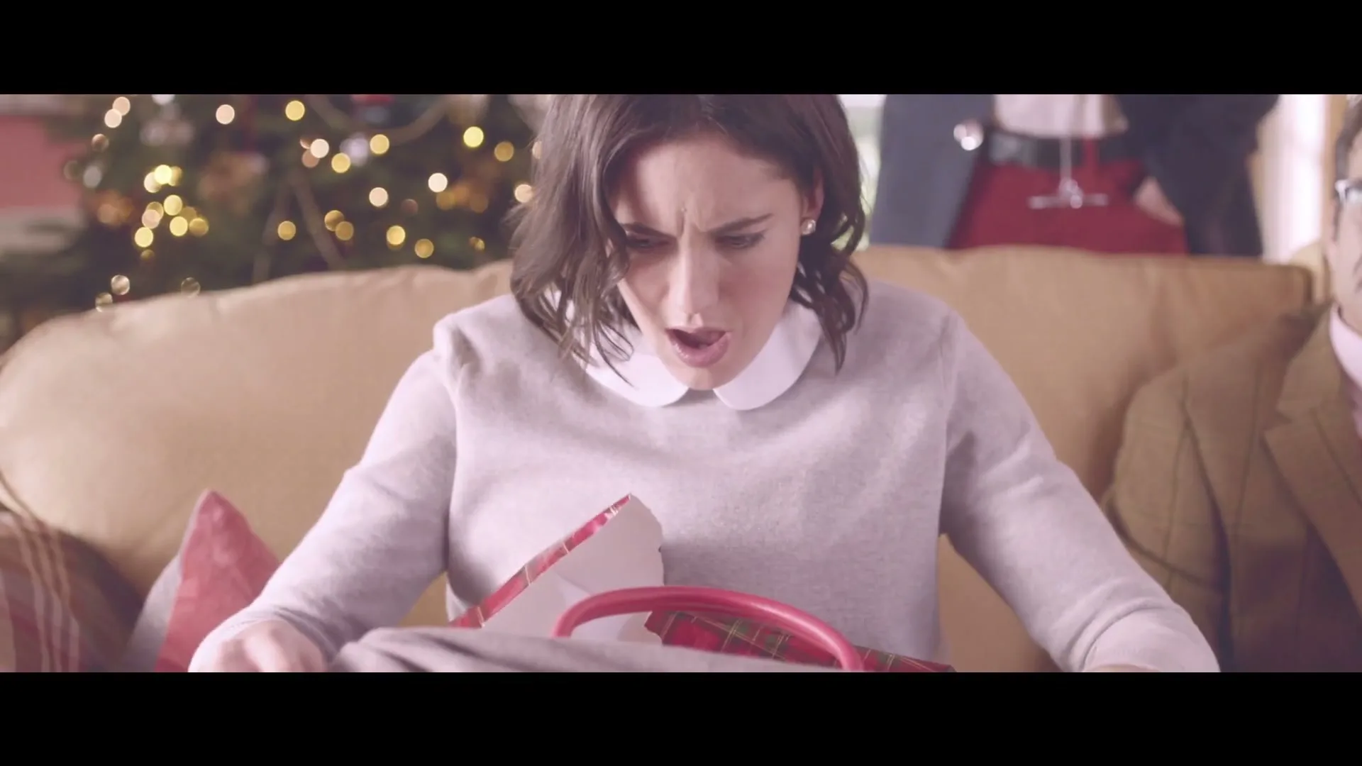 A woman in her 30s, wearing a grey sweater and white collar, opens a gift box to reveal a red handbag, looking surprised and delighted in a festive living room.