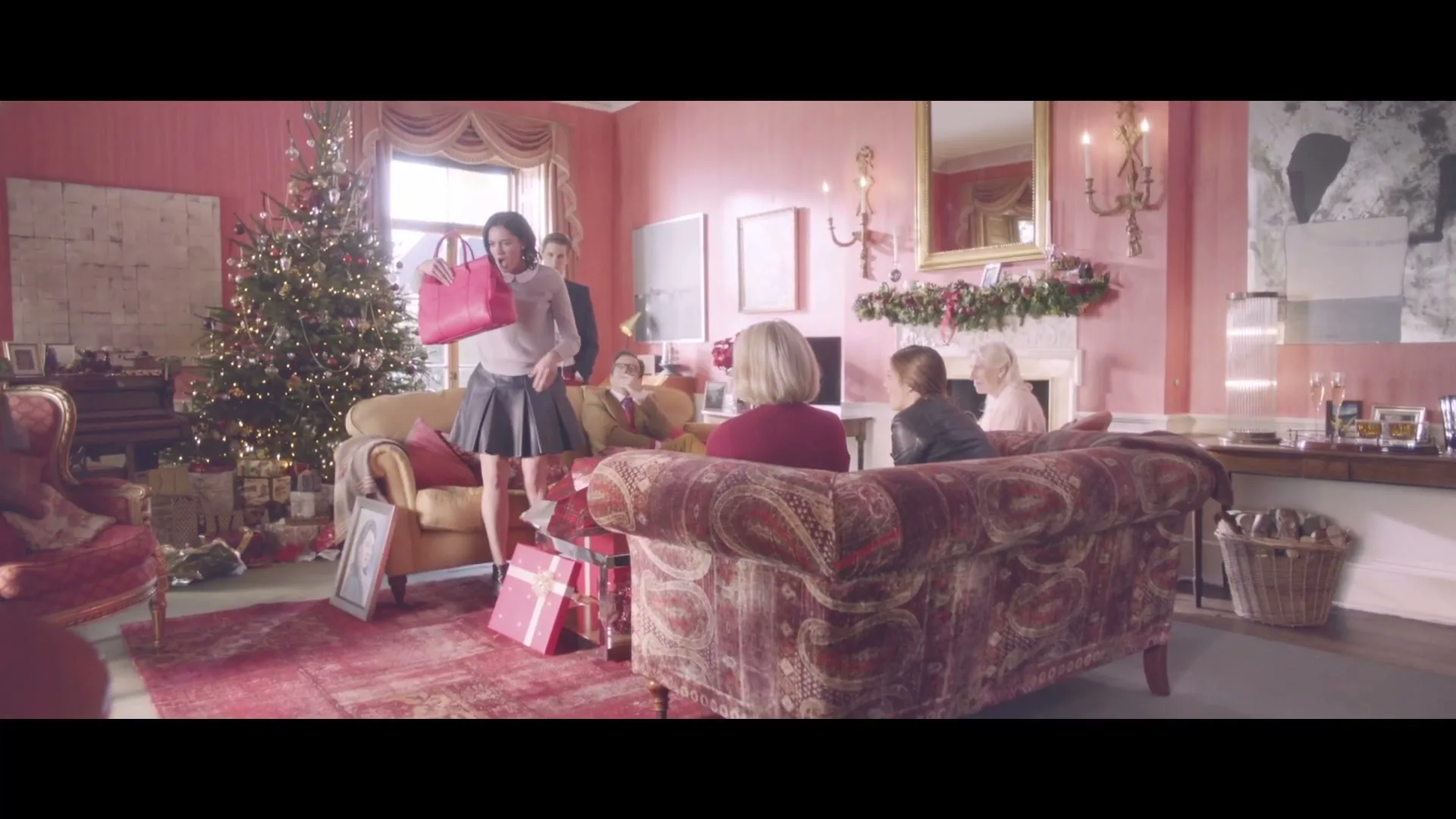 A young woman in a pleated skirt and white sweater holds up a pink handbag, excitedly showing it to her family gathered in a festive living room.